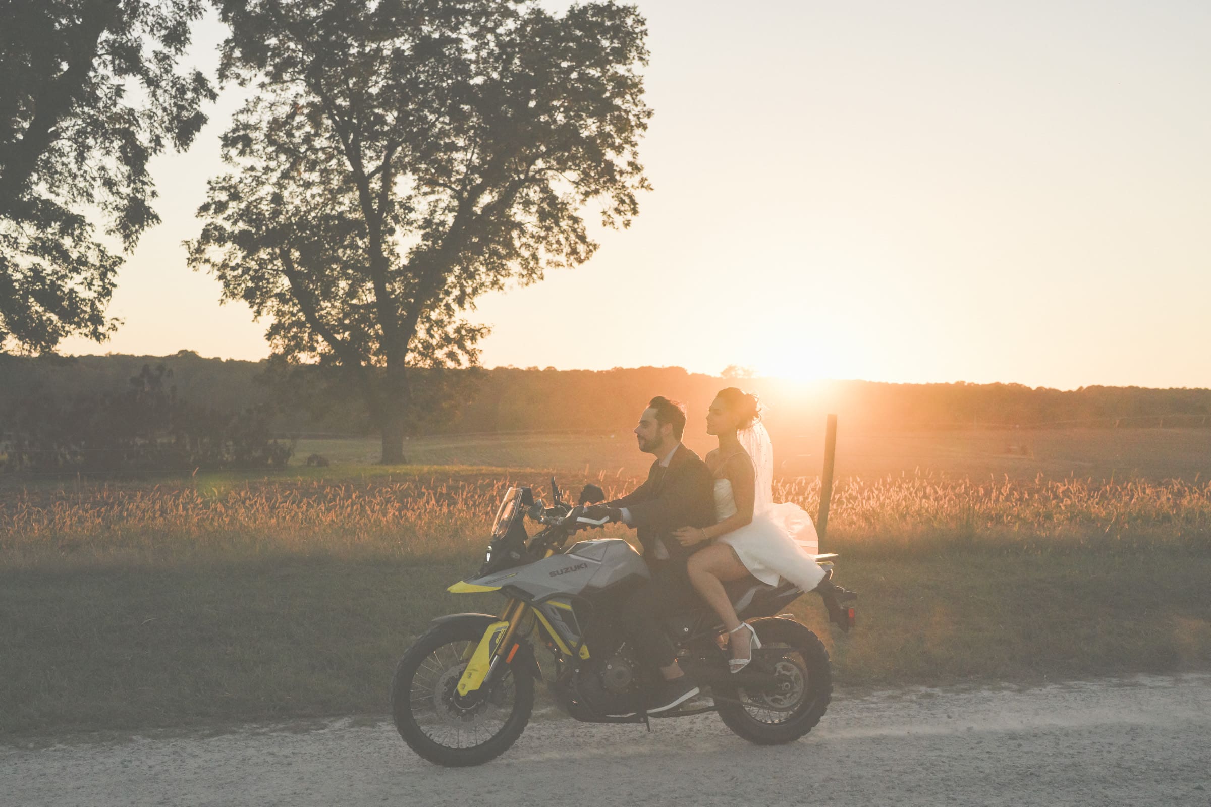 A bride and groom ride a motorcycle on a dirt road at sunset. The groom drives while the bride, in a white dress, sits behind him. The sun sets in the background, casting a warm, golden glow over the scene.
