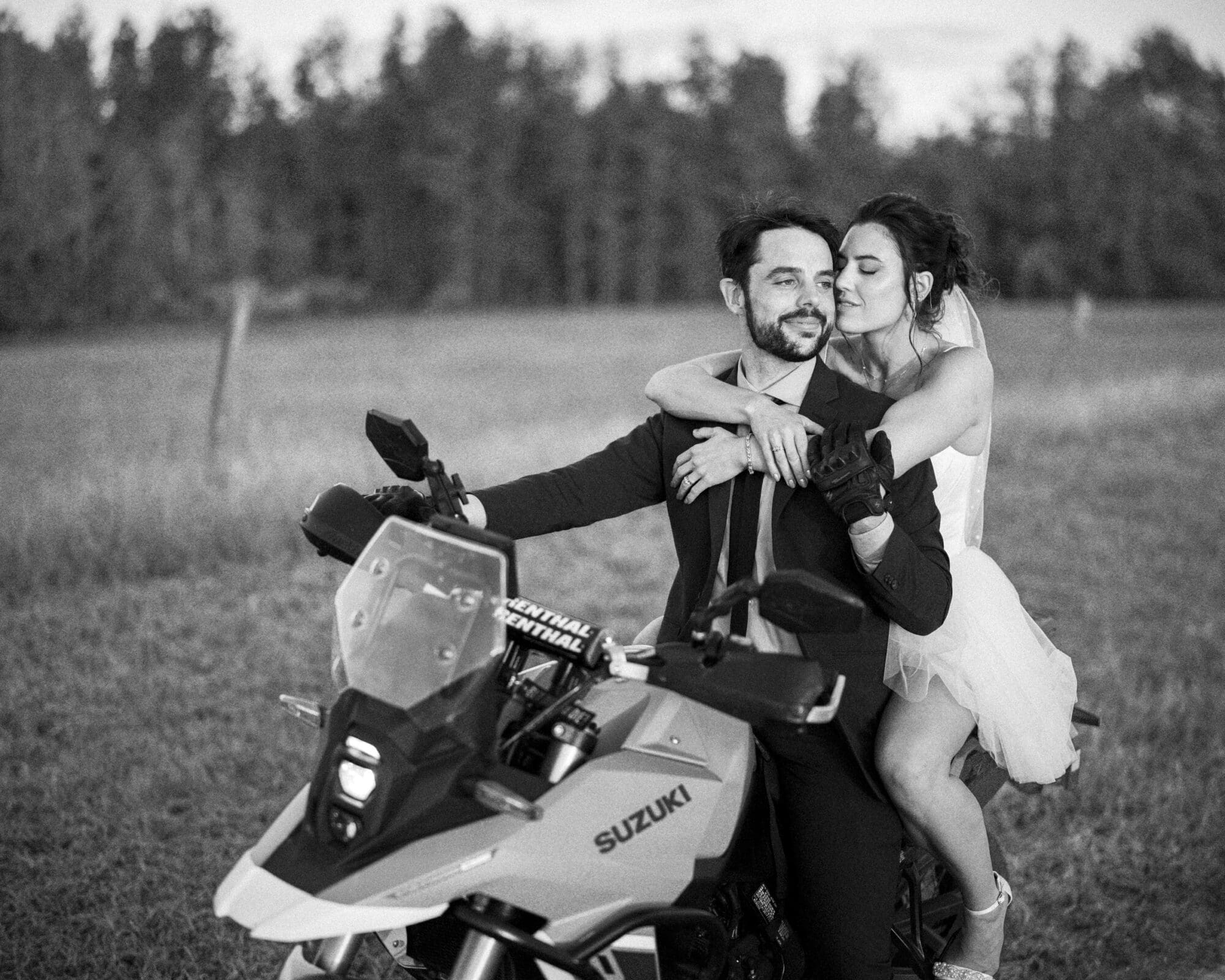 A bride in a white dress hugs a groom in a suit from behind as they sit on a Suzuki motorcycle in a grassy field, sharing a joyful, intimate moment. The background shows trees and an open sky.