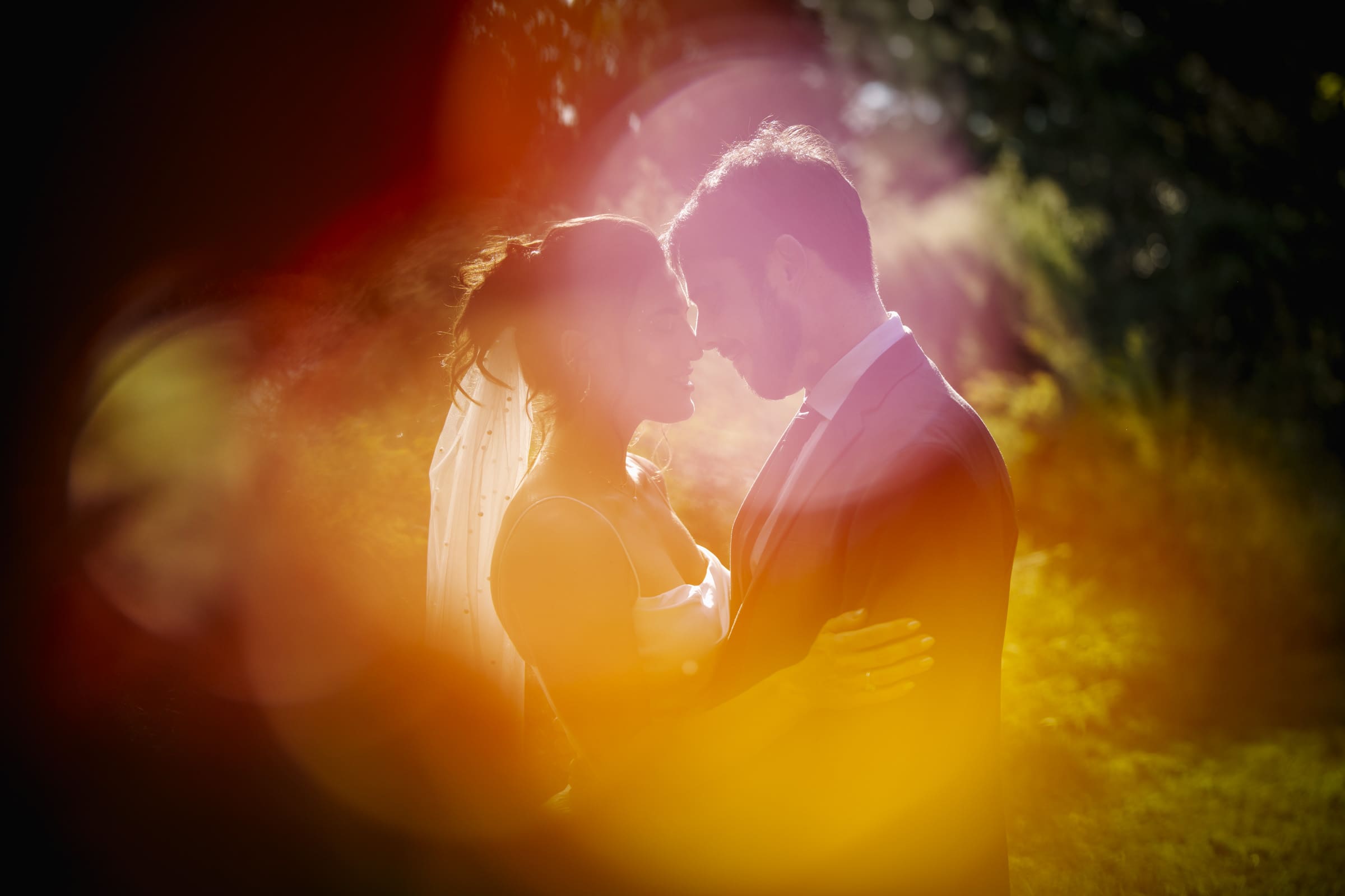 A bride and groom embrace outdoors, touching foreheads and smiling, surrounded by warm, colorful light flares and greenery in the background.