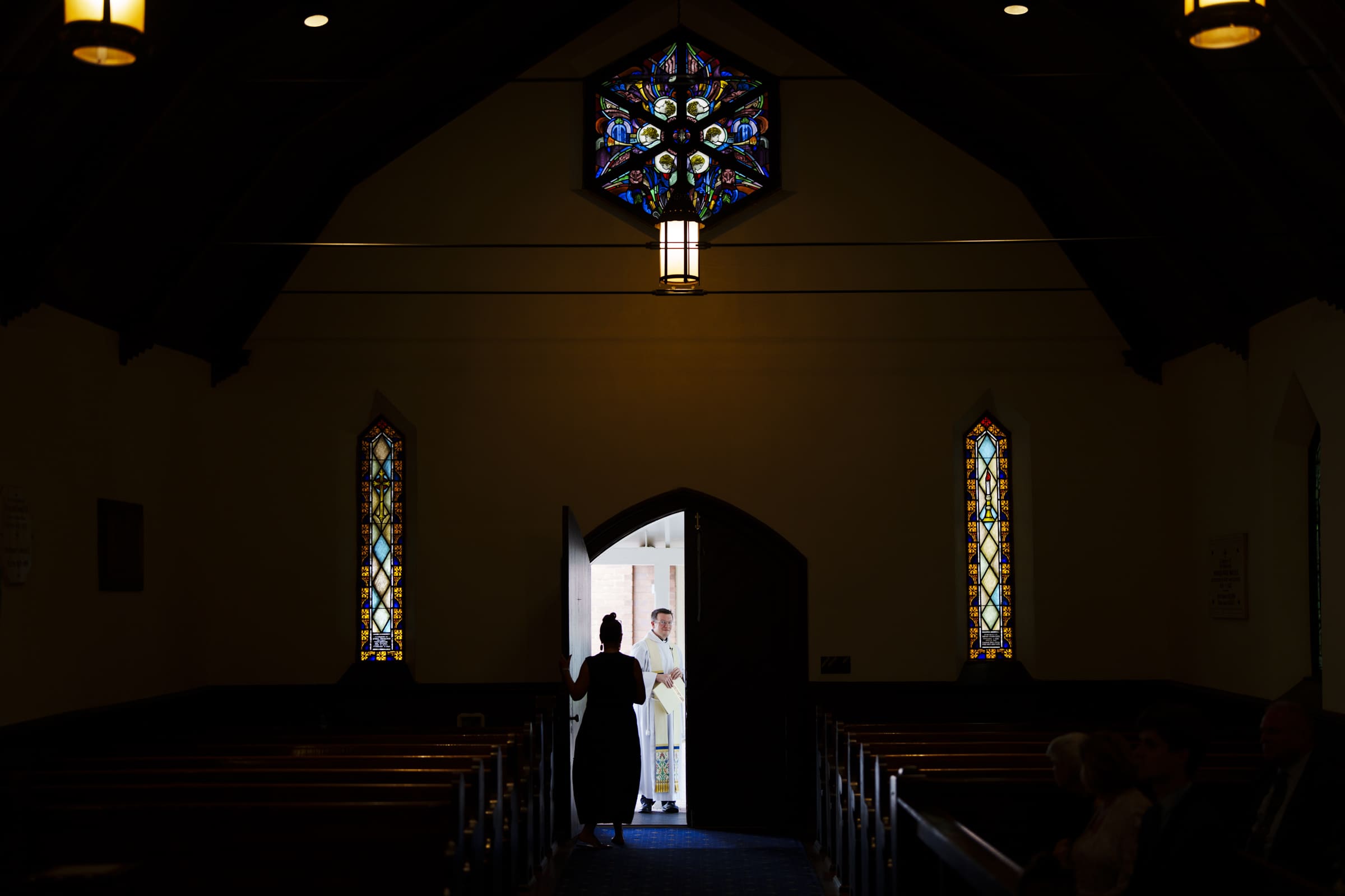 A dimly lit church interior with stained glass windows shows a person standing in the doorway, silhouetted against the light, as another person stands outside in daylight.