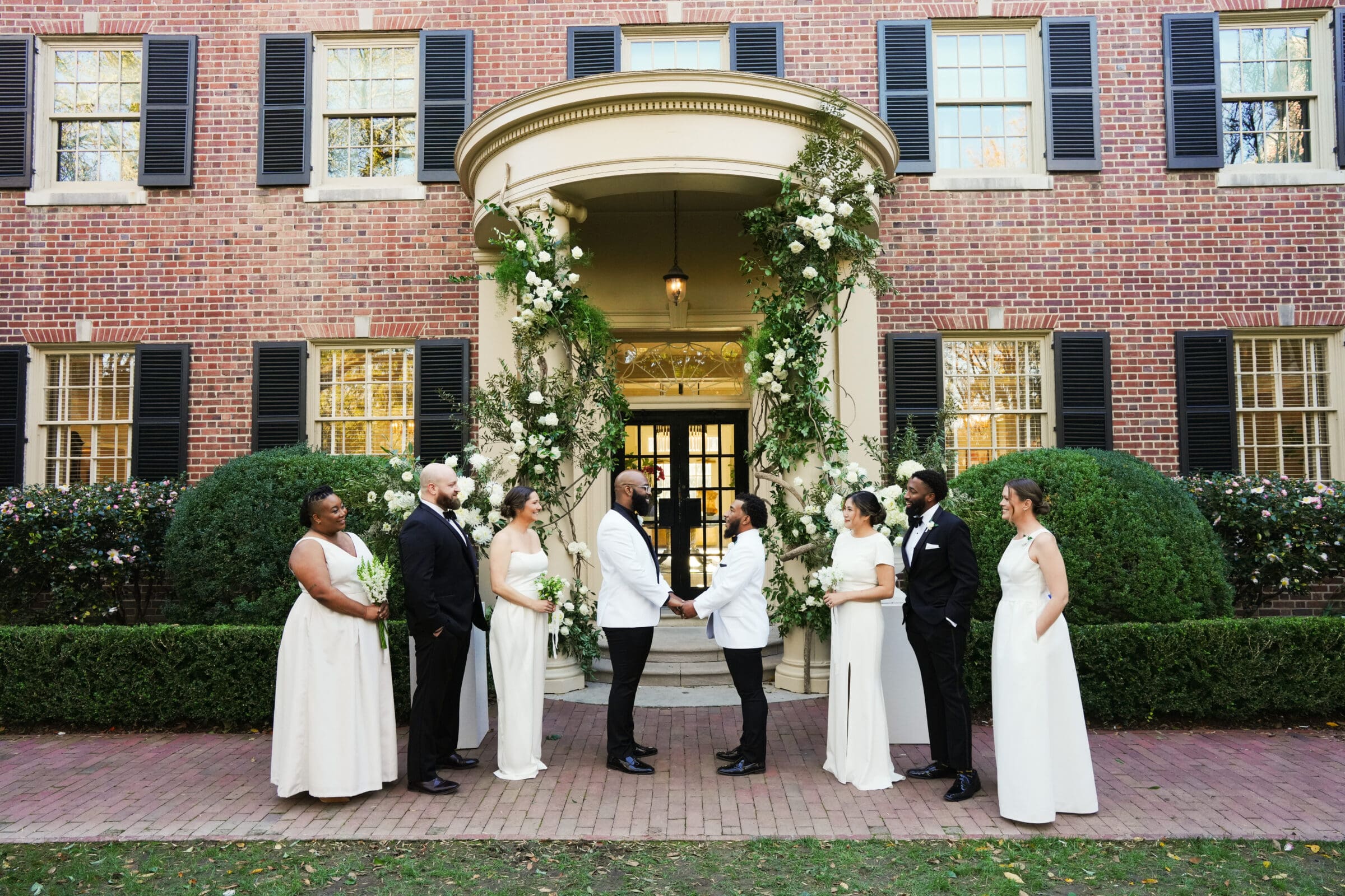 A wedding party poses outside a brick building with black shutters and a floral arch. Two grooms stand together at the center, surrounded by bridesmaids in white and groomsmen in black suits.