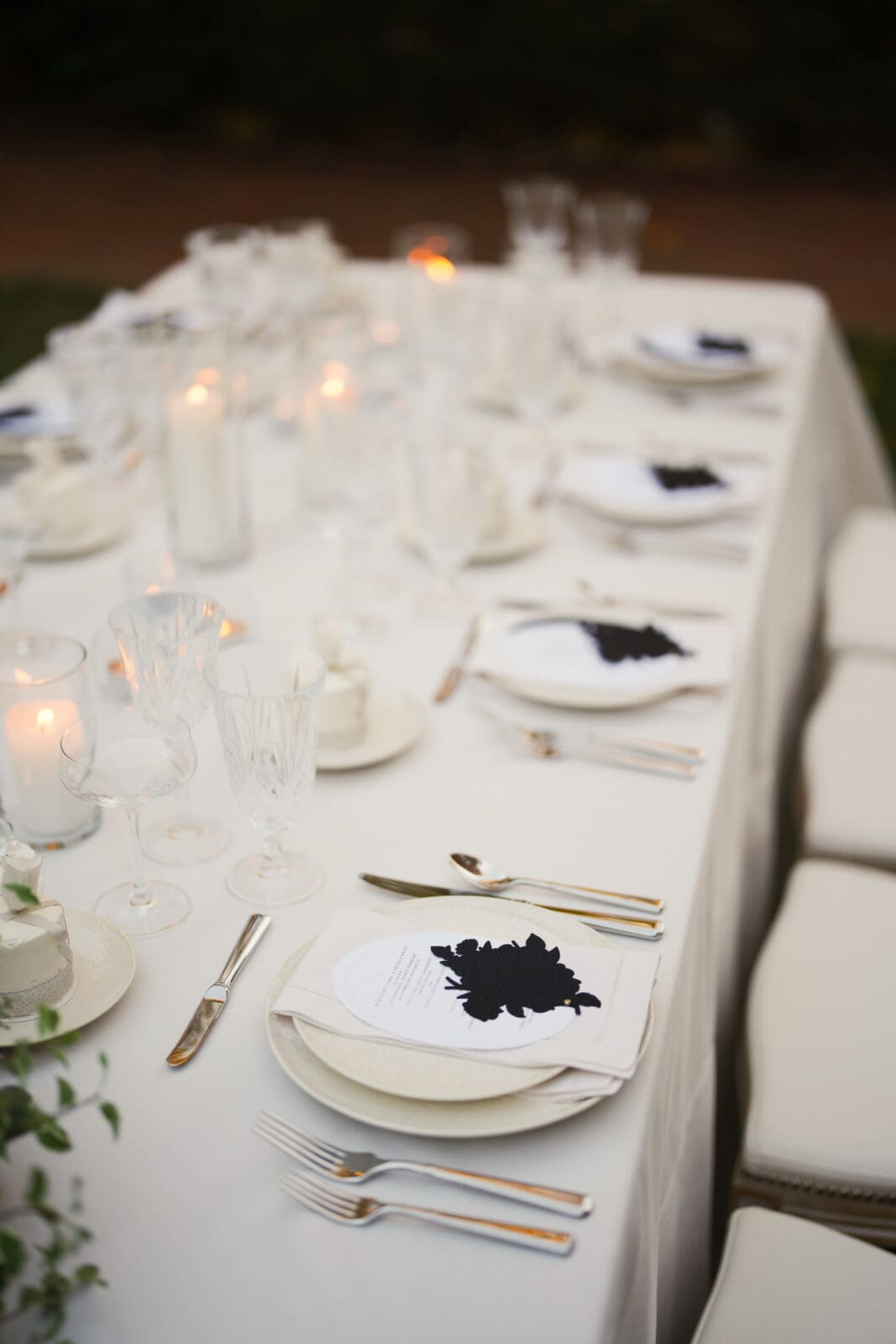 A white table set for a formal event with white plates, silver cutlery, glassware, candles, and menus with a black leaf design. White cushioned chairs line the side of the table.