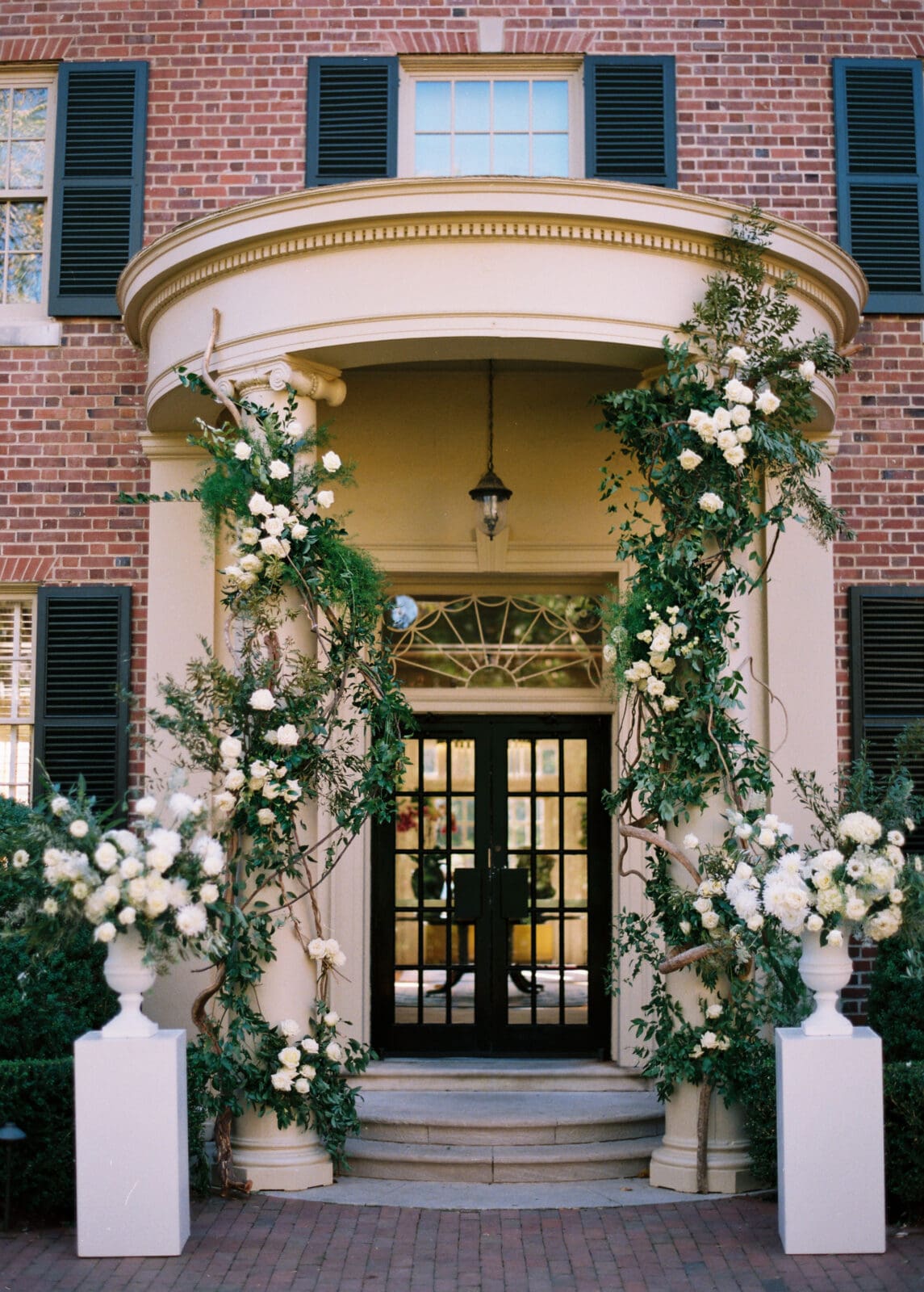 A brick building entrance with black double doors, flanked by two columns decorated with lush greenery and white flowers. Two large white flower arrangements in urns sit on either side of the doorway.