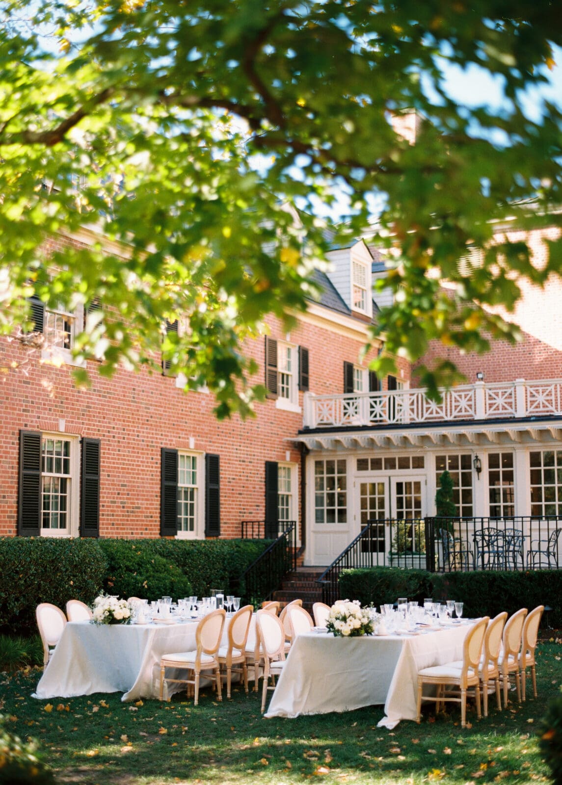 Two elegant tables with white tablecloths and wooden chairs are set for an outdoor meal on a green lawn in front of a large brick house with white trim and dark shutters, under leafy tree branches.