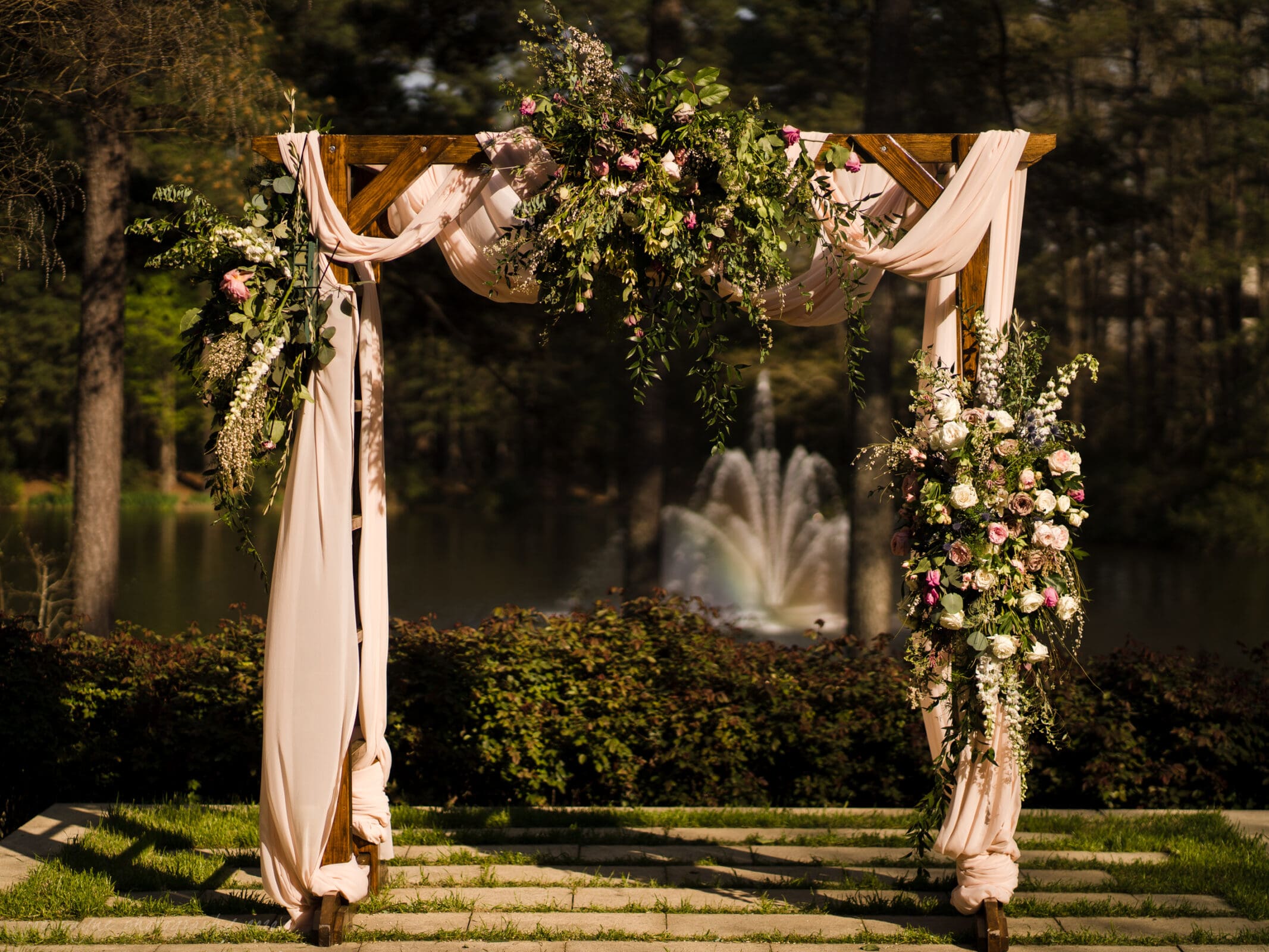 A wooden wedding arch draped with blush pink fabric and decorated with lush green foliage and pink and white flowers, set outdoors with a fountain and trees in the background.