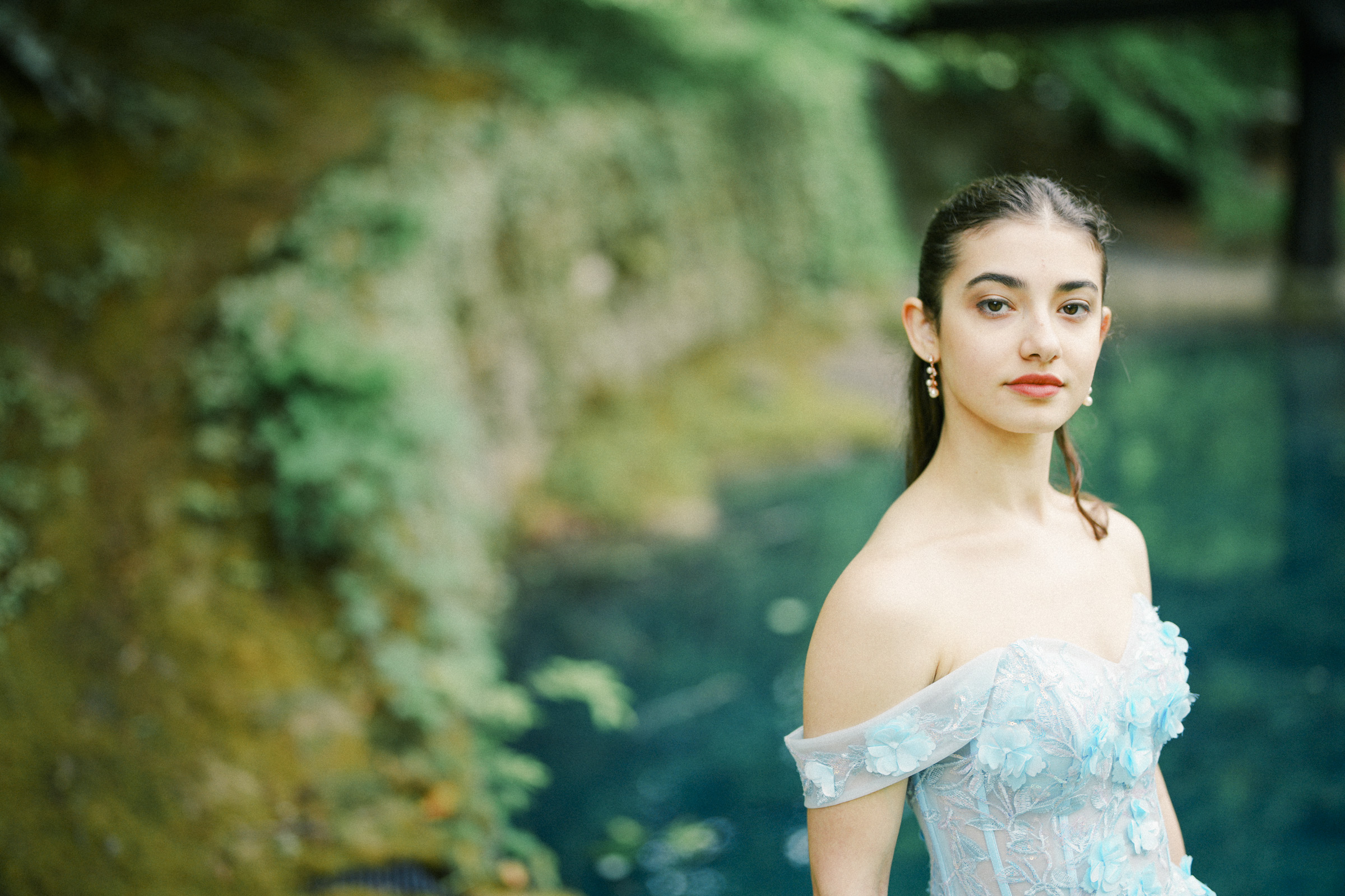 Gianna, dressed in a light blue, off-shoulder floral dress, stands confidently near a lush green area and a blue pond at Castle McCullough, capturing the elegance of her Quinceañera celebration.