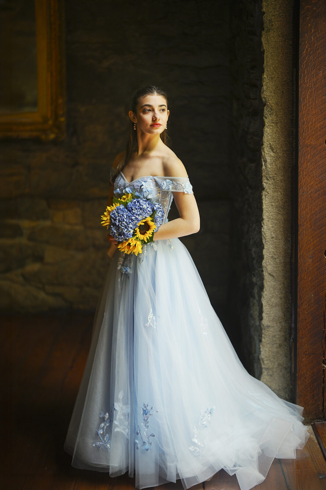 Gianna, dressed in an off-shoulder, light blue Quinceañera gown with floral embroidery, stands by a window at Castle McCullough, holding a bouquet of sunflowers and blue hydrangeas, gazing thoughtfully into the distance.