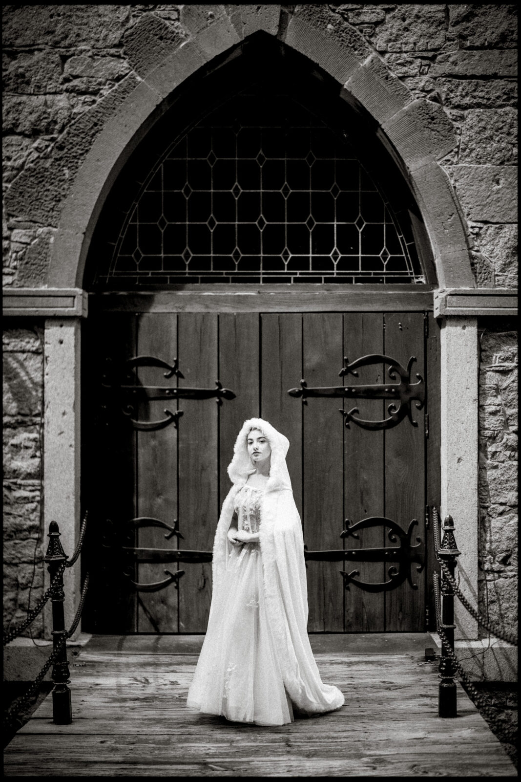 A woman in a white gown and veil stands solemnly before Castle McCullough’s grand church doors. The black-and-white scene evokes a timeless feel, perfect for capturing a Quinceañera Session with vintage elegance.
