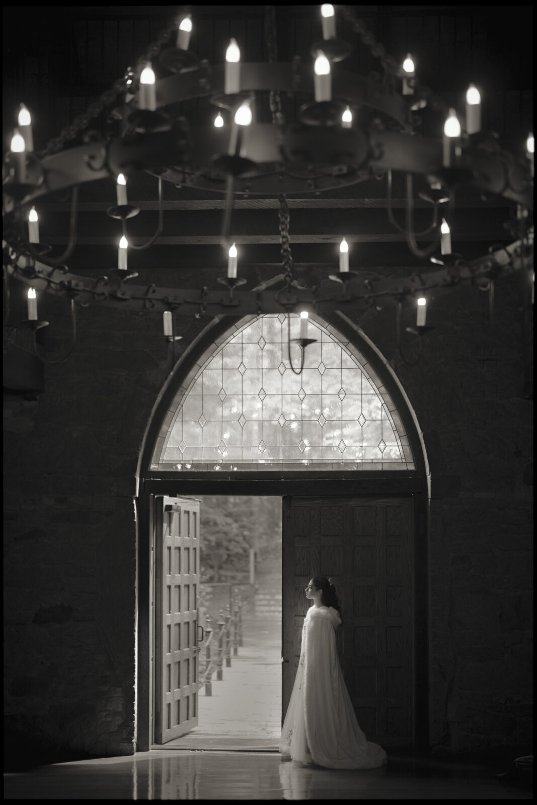 A woman in a long, flowing dress stands by the open door of stone Castle McCullough, its arched window and grand chandelier glowing above—perfect for an enchanting Quinceañera photo session in the dimly lit interior.