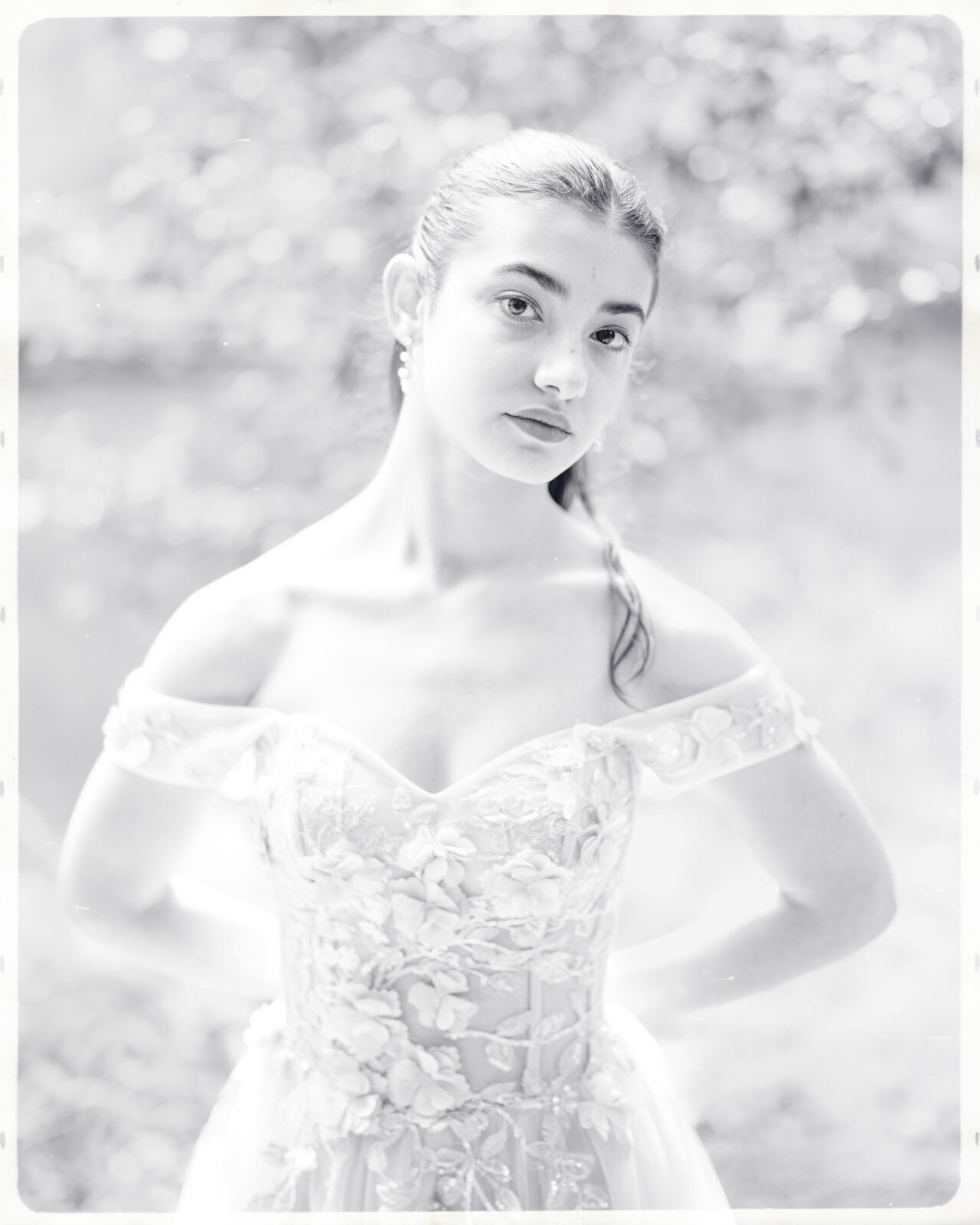 A young woman in an off-the-shoulder, floral-embroidered gown stands outdoors during her Quinceañera Session. The black-and-white photo has a soft, dreamy focus and blurred natural background, evoking fairytale vibes at Castle McCullough.
