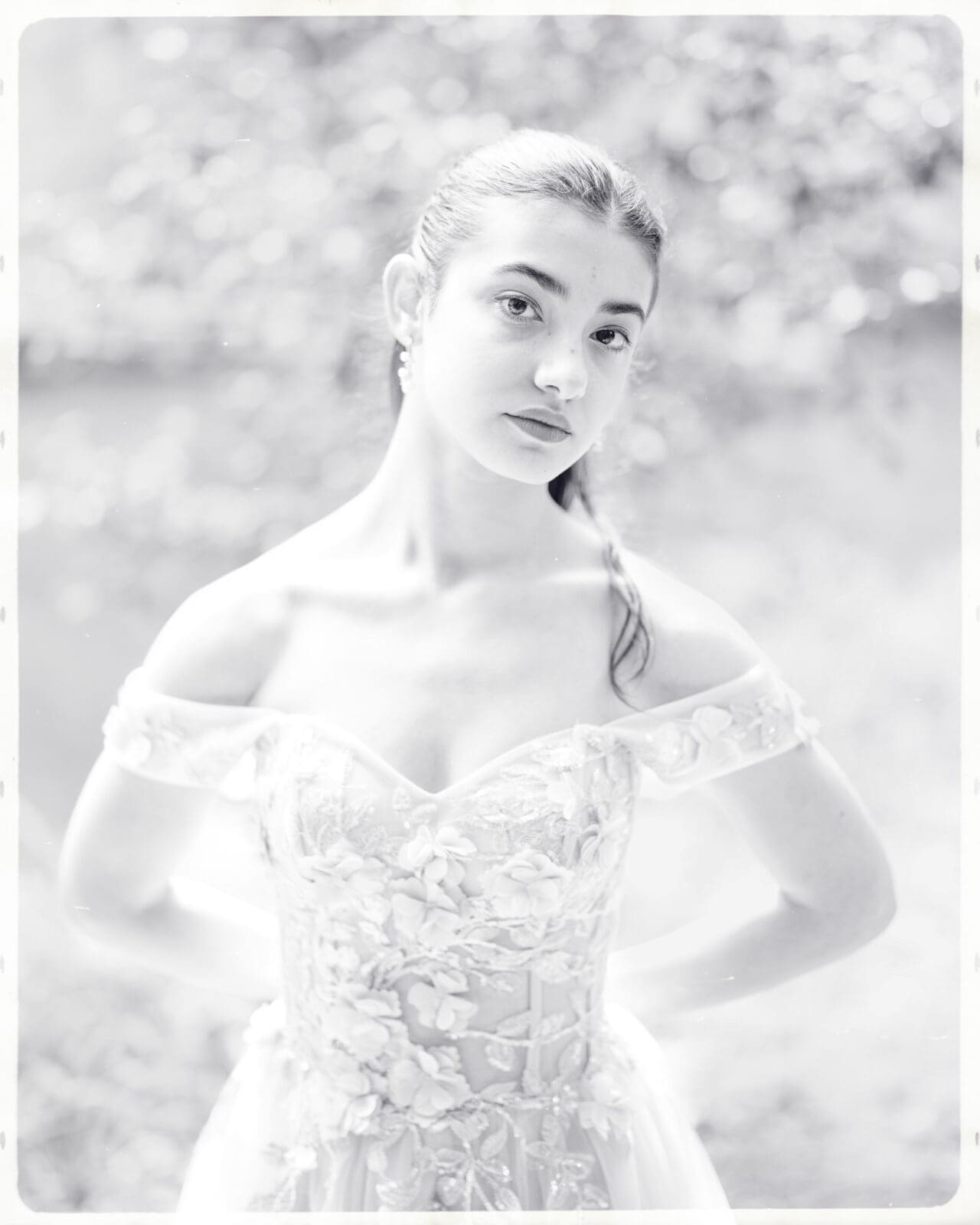 A young woman in an off-the-shoulder, floral-embroidered gown stands outdoors during her Quinceañera Session. The black-and-white photo has a soft, dreamy focus and blurred natural background, evoking fairytale vibes at Castle McCullough.