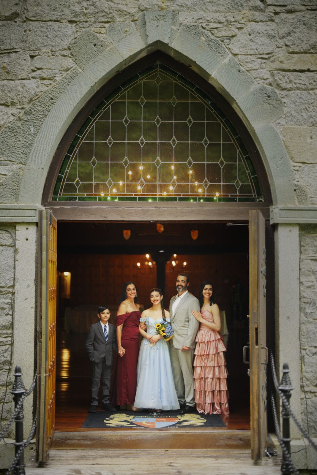 A family dressed in formal attire stands in the open doorway of Castle McCullough, with an arched stained-glass window above. They pose together and smile, celebrating a memorable Quinceañera session.