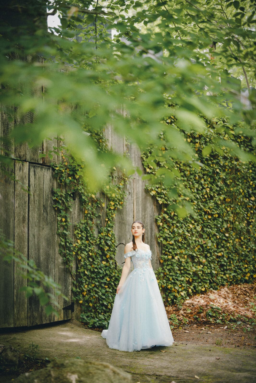 A woman in a light blue, flowing gown stands outdoors near a rustic wooden wall covered in green ivy at Castle McCullough, surrounded by lush greenery and fallen leaves—perfect for a magical Quinceañera session.