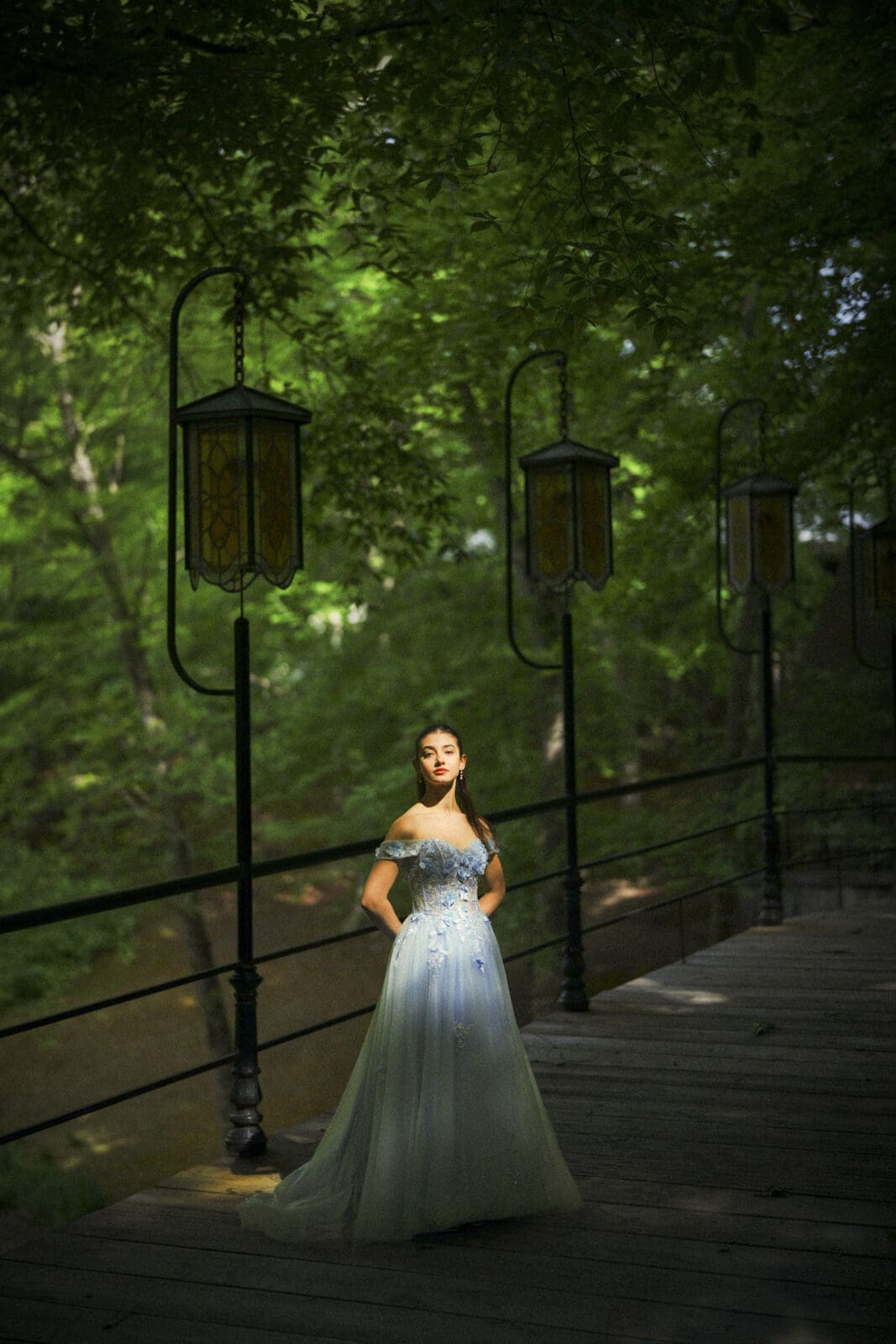 Gianna, celebrating her Quinceañera, stands on a wooden bridge at Castle McCullough in a light blue off-shoulder gown, surrounded by lush green trees and vintage lanterns glowing in the sunlight.