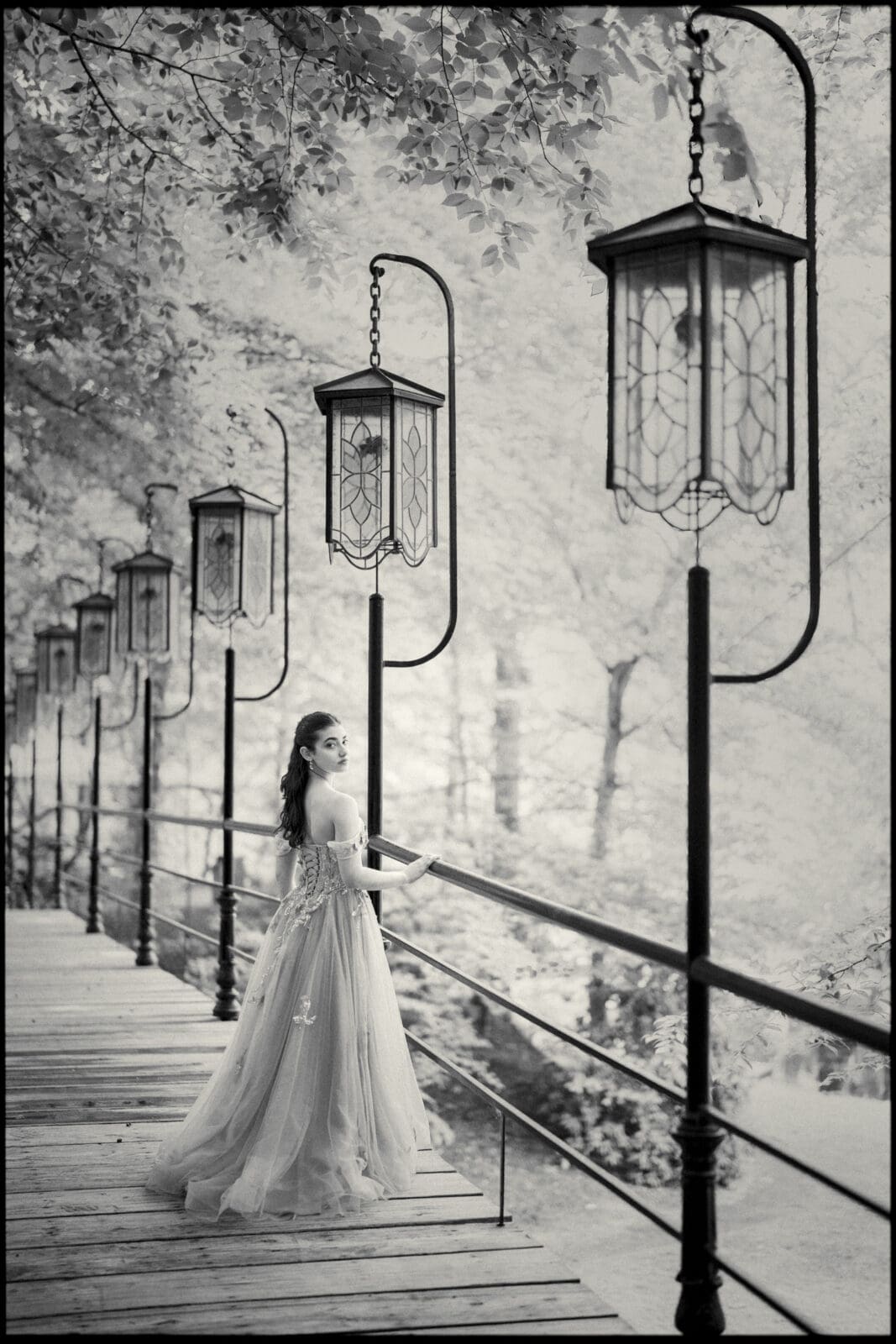 A woman in a flowing, elegant gown stands on a wooden bridge lined with tall lanterns at Castle McCullough, surrounded by dense trees and soft light, creating a dreamy, ethereal Quinceañera Session atmosphere.