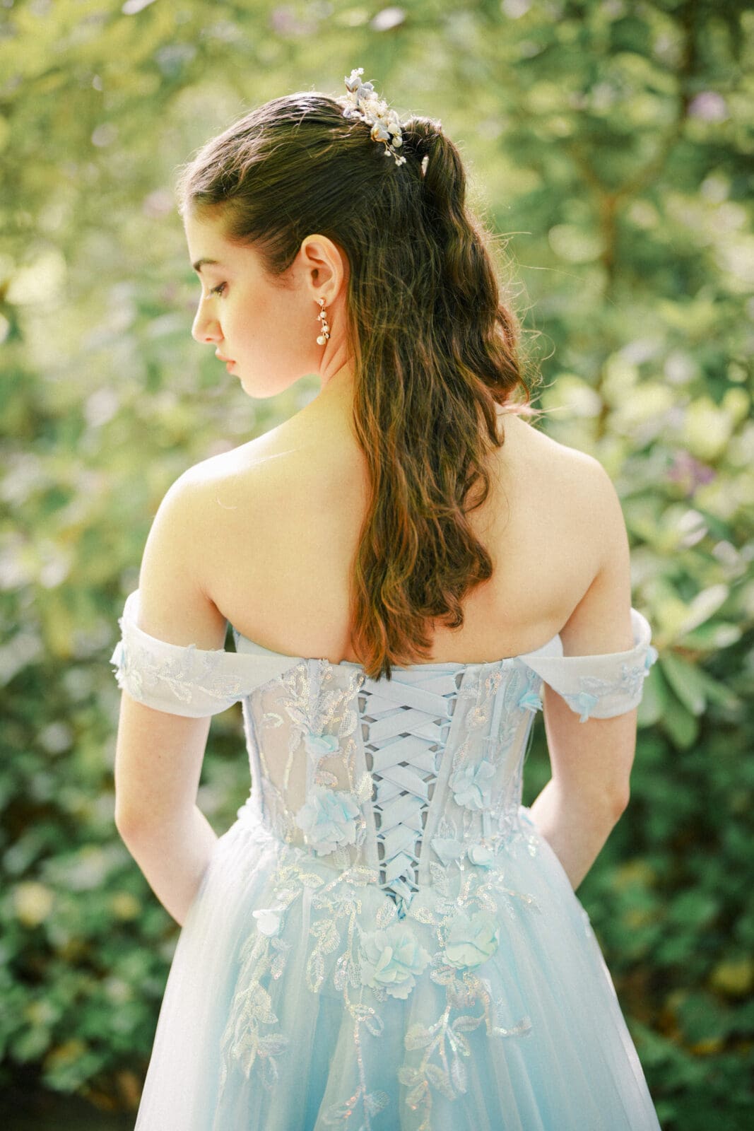A young woman in a light blue, off-the-shoulder gown with floral details stands outdoors at Castle McCullough for her Quinceañera session. Her long brown hair is half up, adorned with a decorative hairpiece, surrounded by lush greenery.