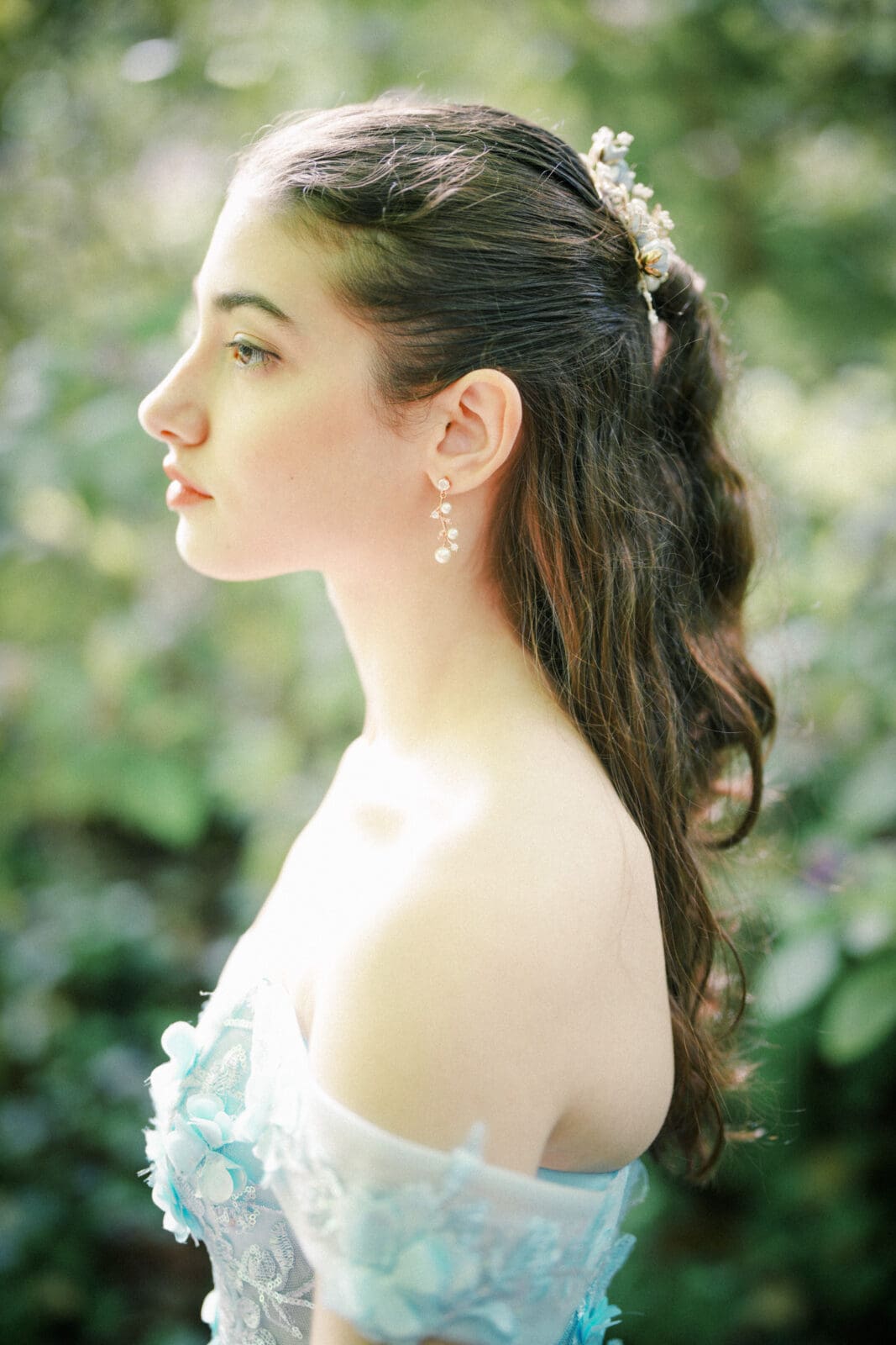 A young woman with long brown hair, styled with a floral hair accessory, stands in profile outdoors during her Quinceañera Session at Castle McCullough. She wears a light blue off-the-shoulder dress with floral details and drop earrings, set against a soft green background.