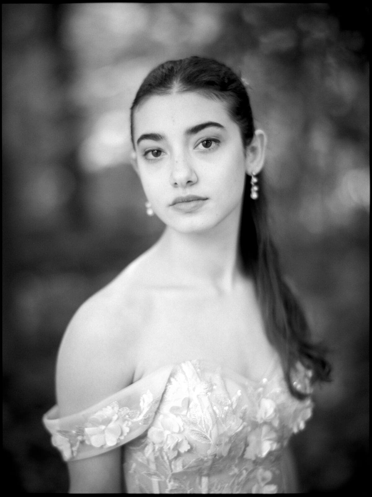 A young woman with long dark hair, wearing an off-the-shoulder, floral-patterned dress and drop earrings, gazes softly at the camera during a photography session. The black-and-white photo features a blurred, natural background.