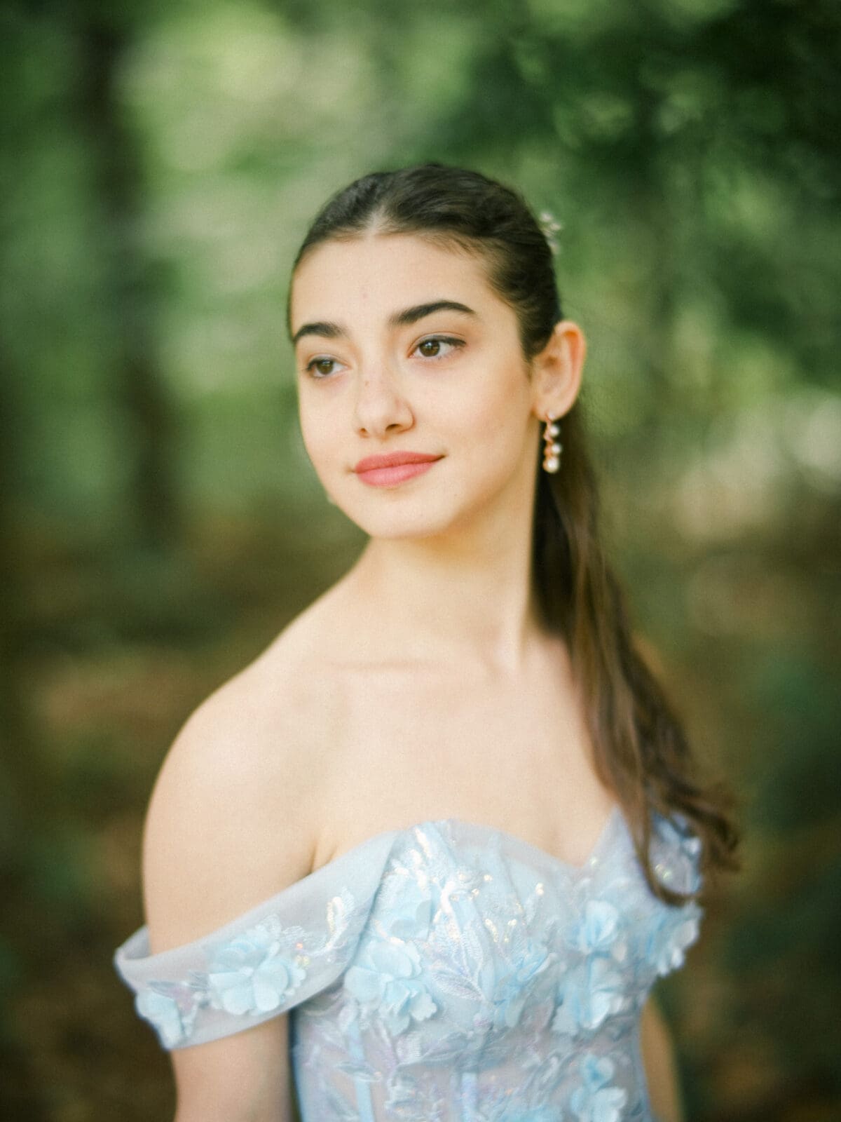 Gianna, dressed in a light blue off-shoulder dress with floral details and drop earrings, stands outdoors at Castle McCullough, the soft, blurred green background highlighting her Quinceañera elegance.