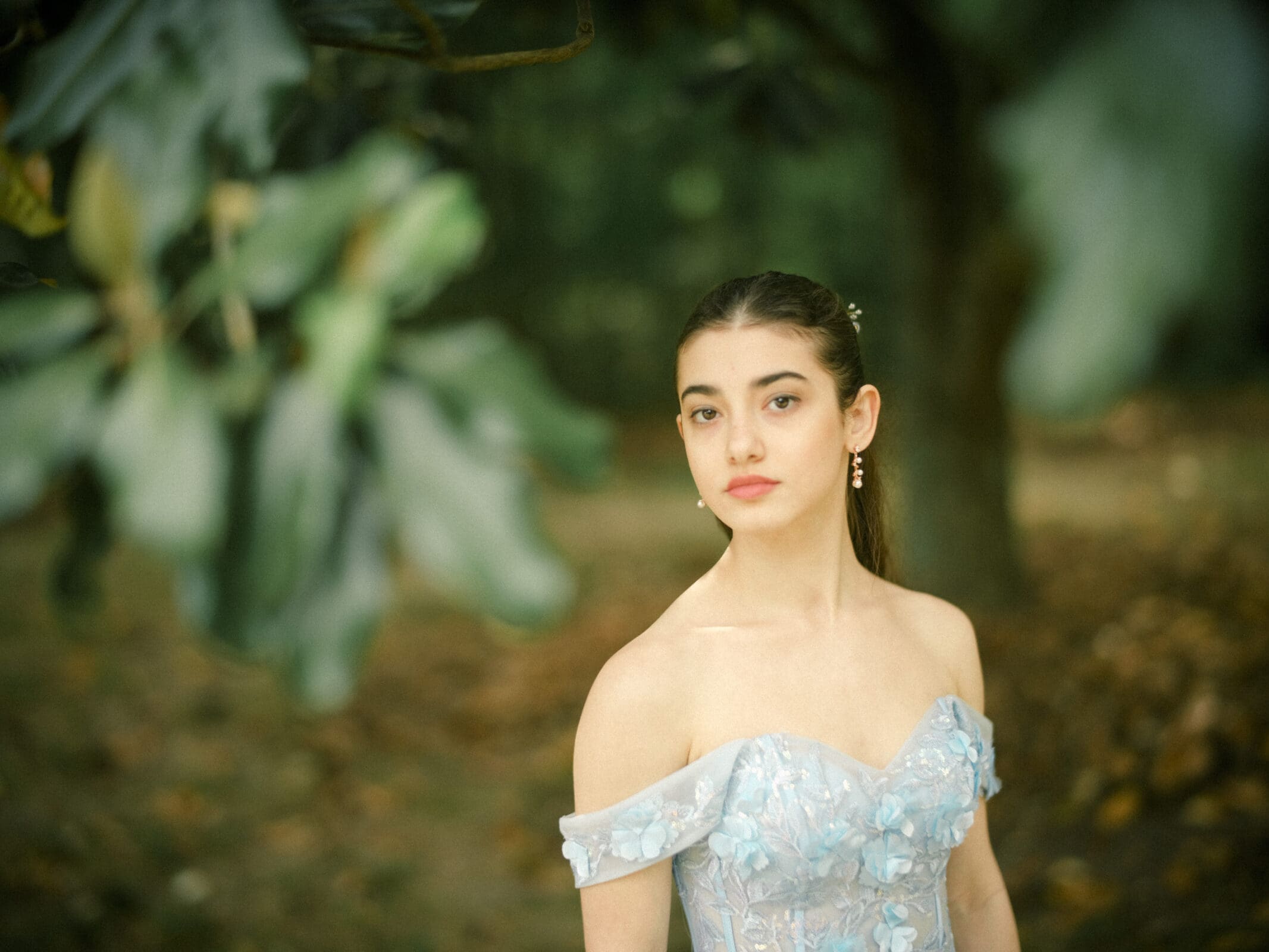 A young woman in an off-the-shoulder, light blue floral dress stands outdoors at Castle McCullough, looking at the camera with a calm expression. The blurred greenery and trees set the perfect backdrop for her Quinceañera session.