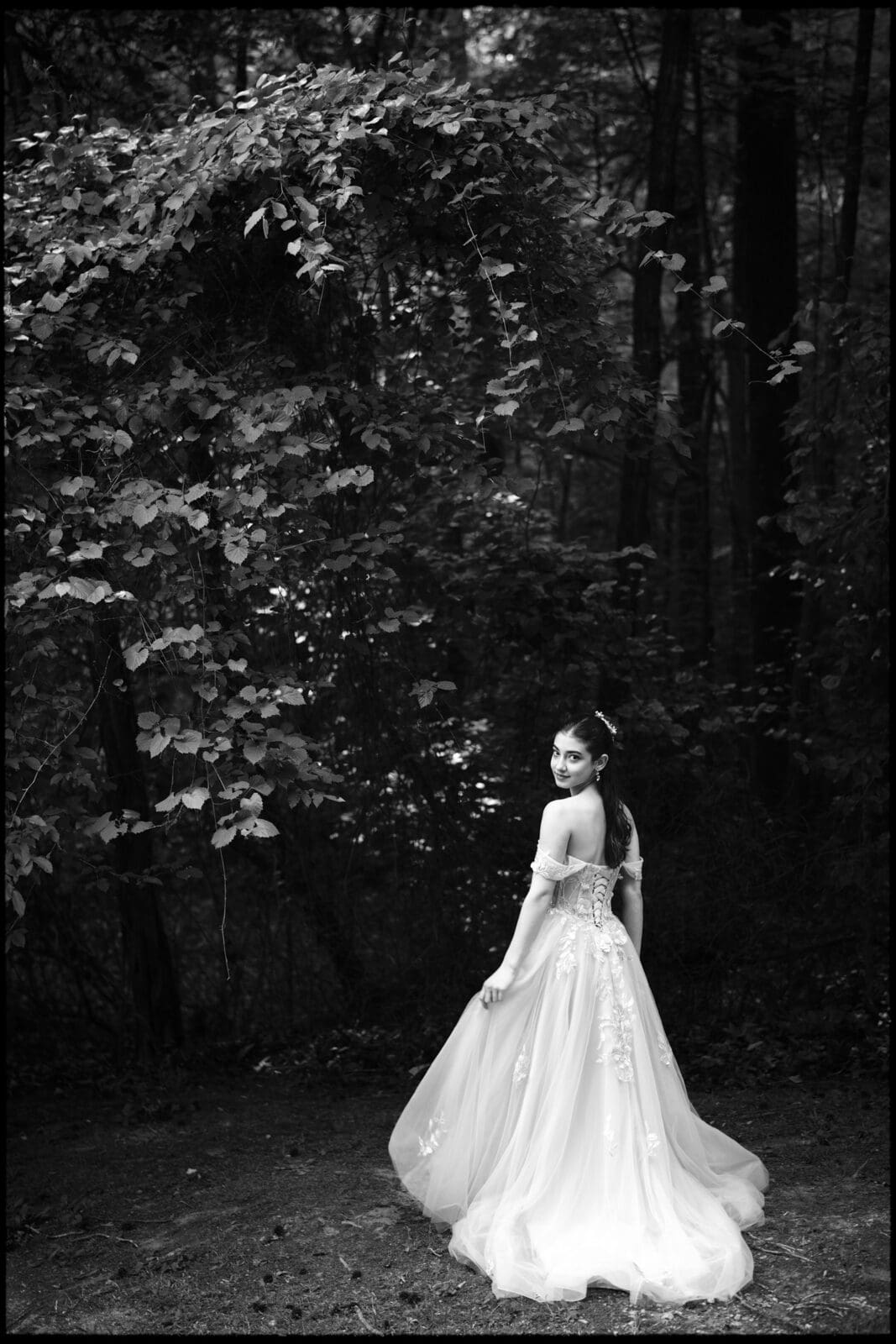 A woman in an elegant, flowing gown stands in a forest, looking over her shoulder toward the camera. Sunlight filters through dense foliage, creating a serene, fairy-tale atmosphere in this black and white Quinceañera Session.