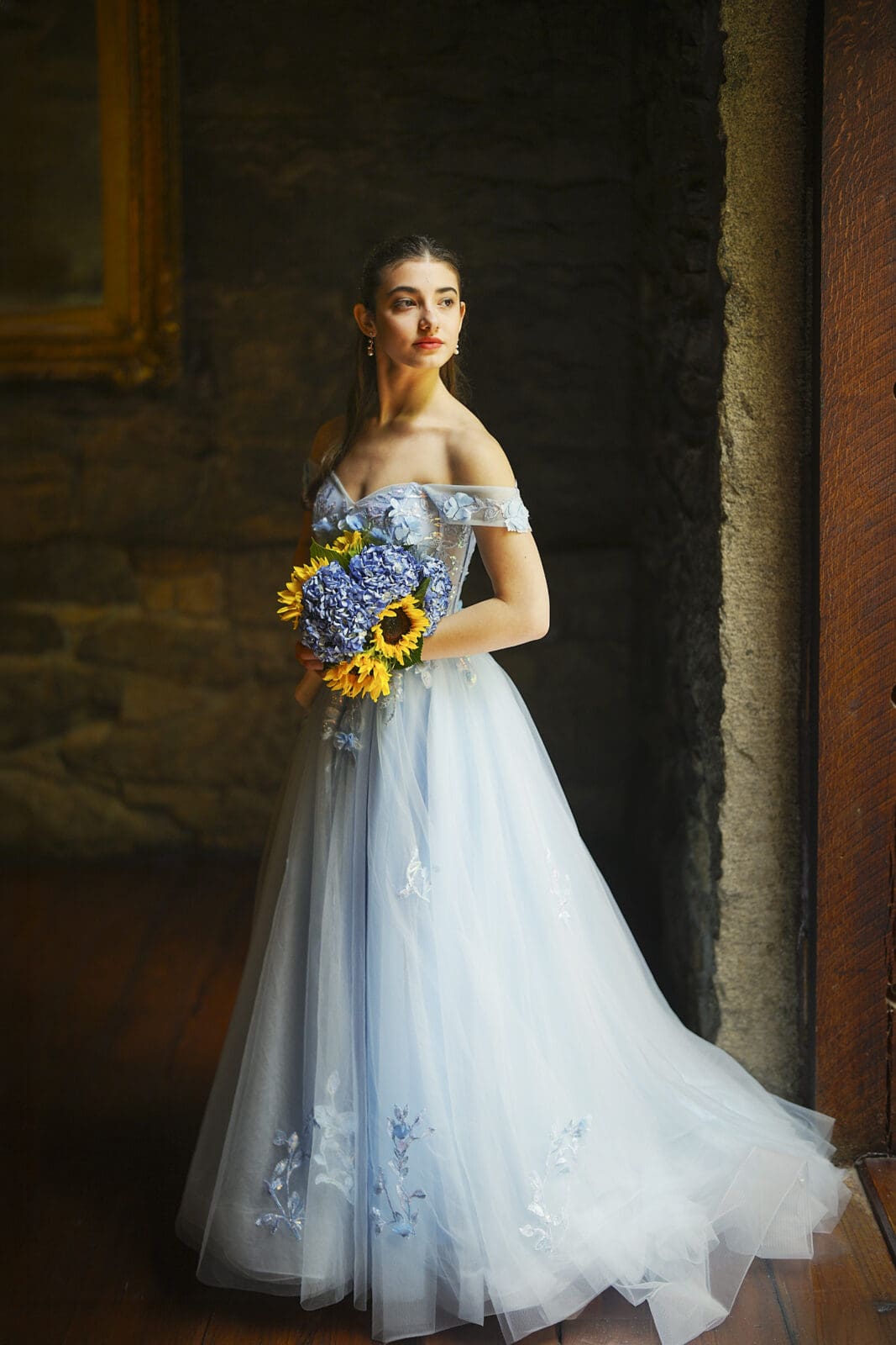 Gianna, dressed in an off-shoulder, light blue Quinceañera gown with floral embroidery, stands by a window at Castle McCullough, holding a bouquet of sunflowers and blue hydrangeas, gazing thoughtfully into the distance.