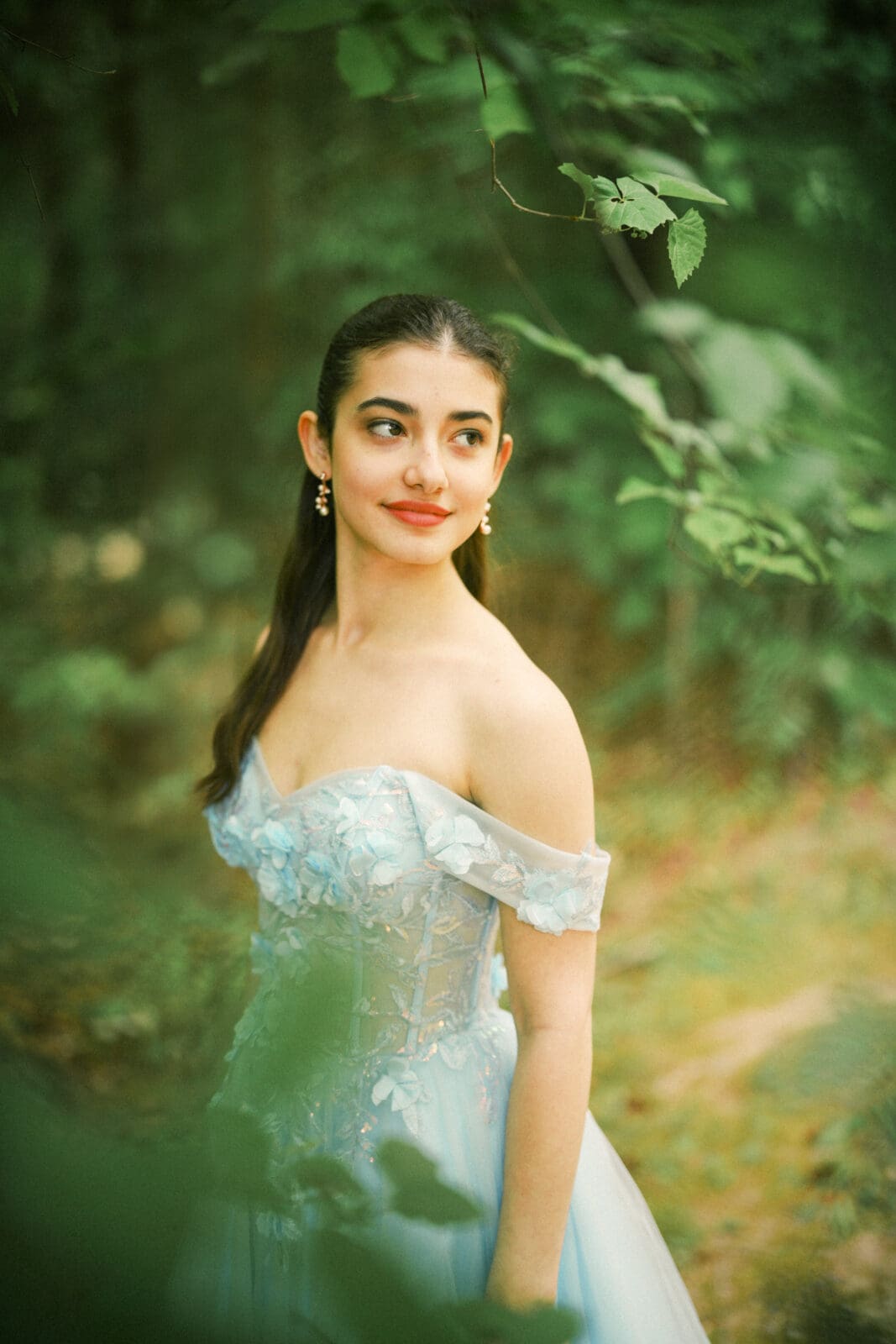 A woman in a light blue off-shoulder gown stands in a lush green forest during her Quinceañera photography session, looking to the side with a gentle smile as sunlight filters through the leaves, creating a soft and dreamy atmosphere.