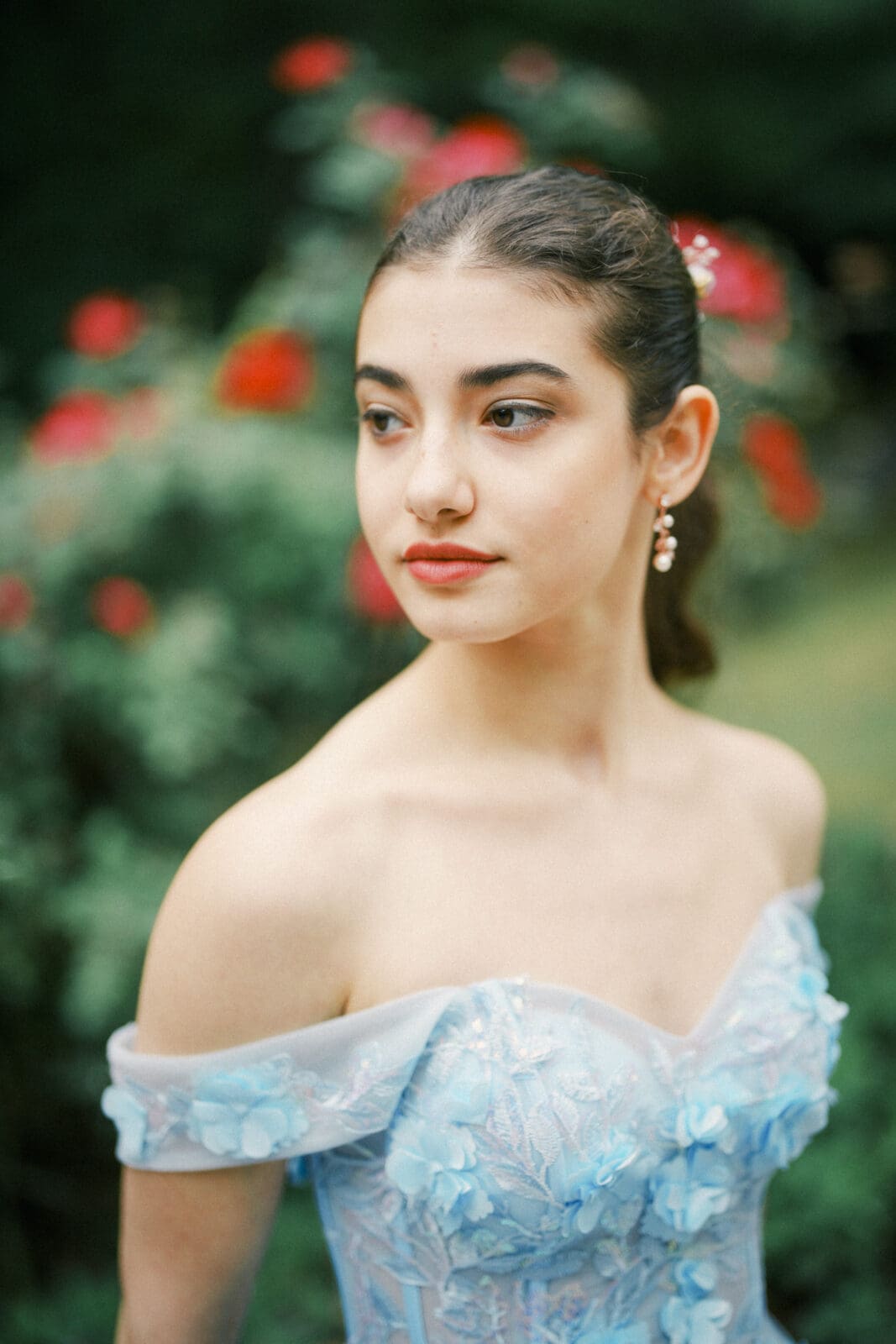 Gianna, dressed in an off-the-shoulder blue dress with floral details, stands outside Castle McCullough, looking to the side. Her hair is tied back as she poses among blurred red flowers and greenery, ready for her Quinceañera celebration.