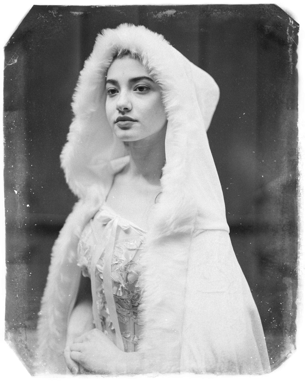 A young woman in a light, fur-trimmed hooded cloak and a lace-up dress poses for a vintage-style Quinceañera portrait at Castle McCullough, gazing slightly to the side with a calm expression. The photo has an old, worn effect.