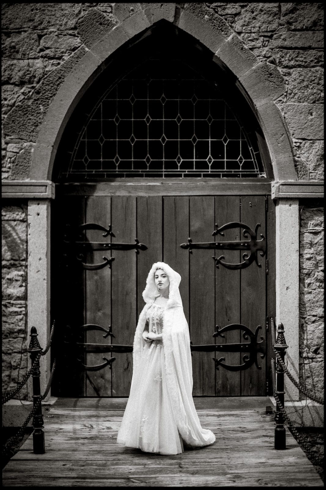 A woman in a white gown and veil stands solemnly before Castle McCullough’s grand church doors. The black-and-white scene evokes a timeless feel, perfect for capturing a Quinceañera Session with vintage elegance.