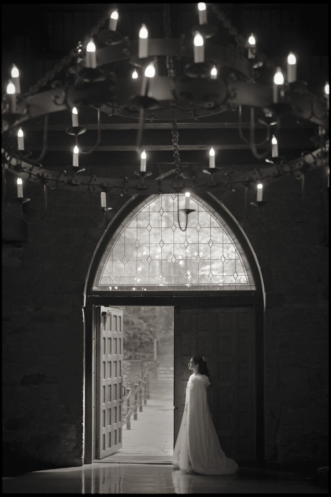 A woman in a long, flowing dress stands by the open door of stone Castle McCullough, its arched window and grand chandelier glowing above—perfect for an enchanting Quinceañera photo session in the dimly lit interior.