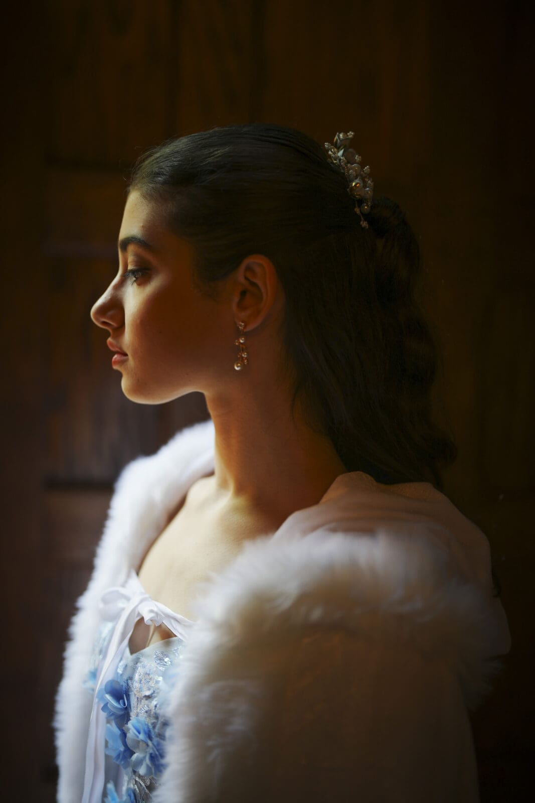 A young woman with long dark hair, wearing a white fur-trimmed garment and pearl earrings, is shown in profile against a dark wooden background at Castle McCullough, softly lit from the side during her Quinceañera Session.