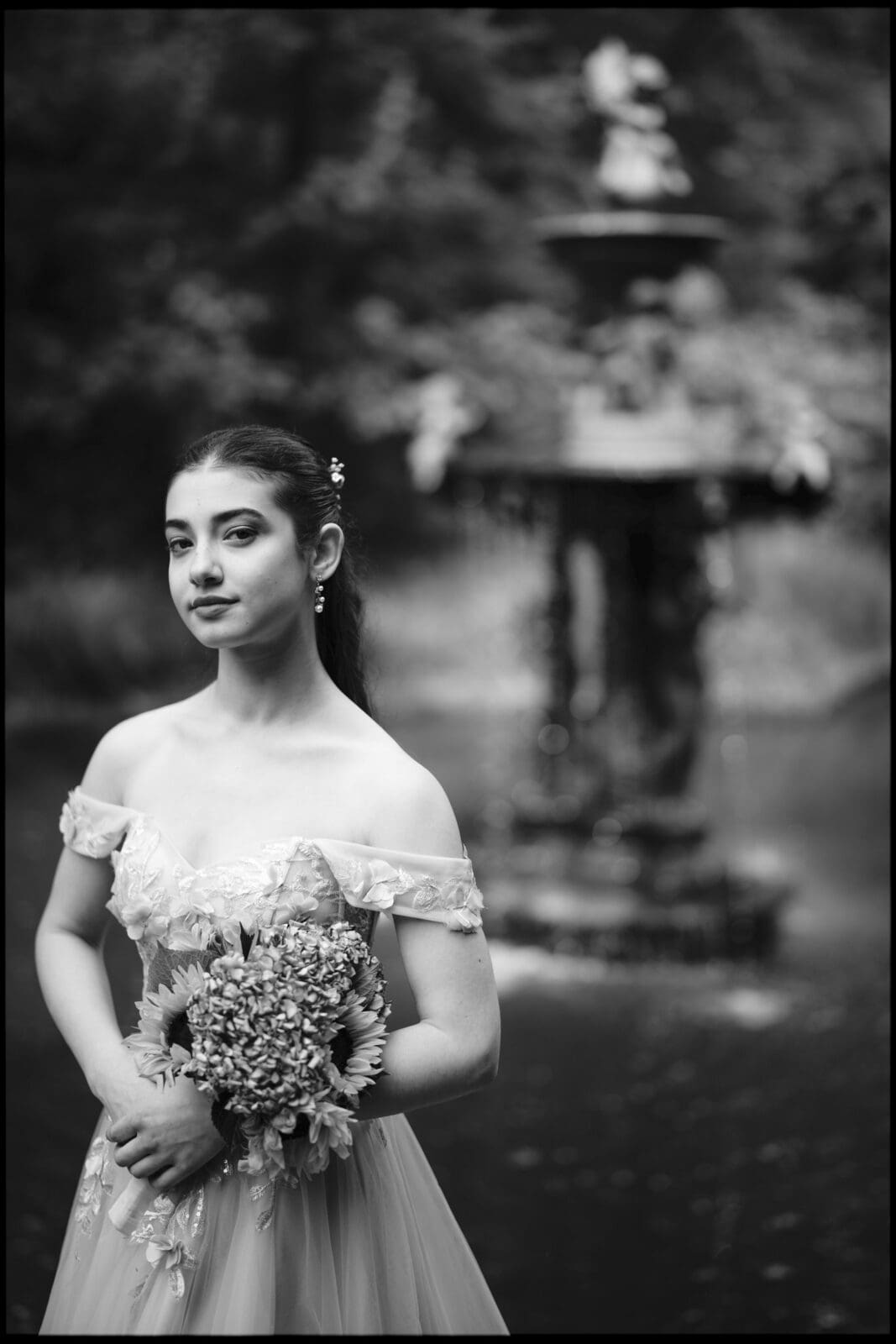 A young woman in an off-shoulder dress holds a bouquet of flowers and stands calmly before a decorative fountain in a park, captured during her Quinceañera Session. The elegant black-and-white image echoes the timeless beauty of Castle McCullough.