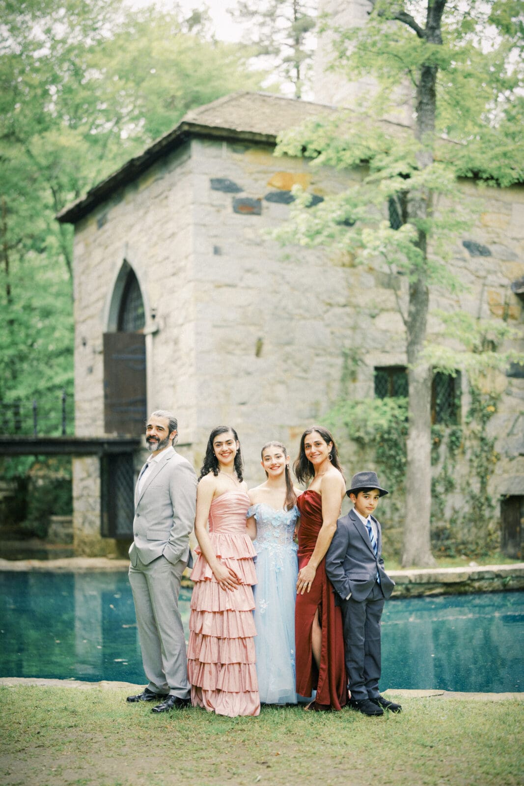 A family of five, dressed formally, stands in front of Castle McCullough beside a reflective pond. The group includes an adult man, three women in colorful dresses—perfect for a Quinceañera session—and a young boy in a suit and hat. Trees surround the scene.