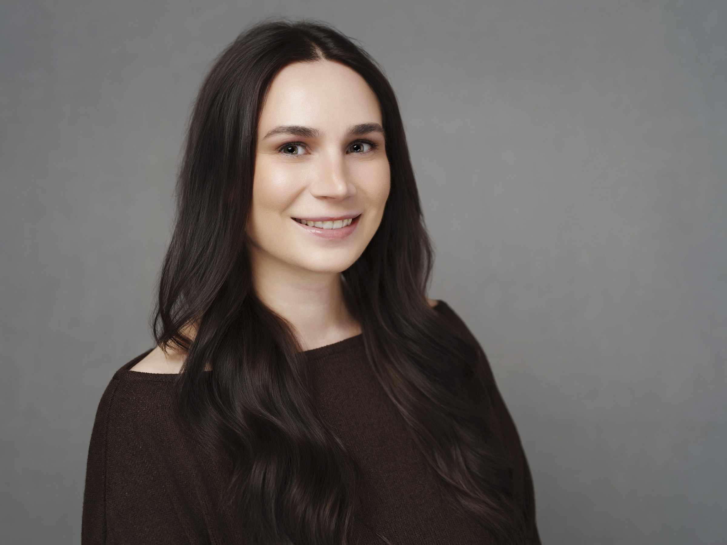 A woman with long, dark brown hair and light skin smiles at the camera in a classic corporate headshot. She wears a brown top and is posed against a plain gray background, perfect for professional profiles at The Umstead Hotel or Macrae.