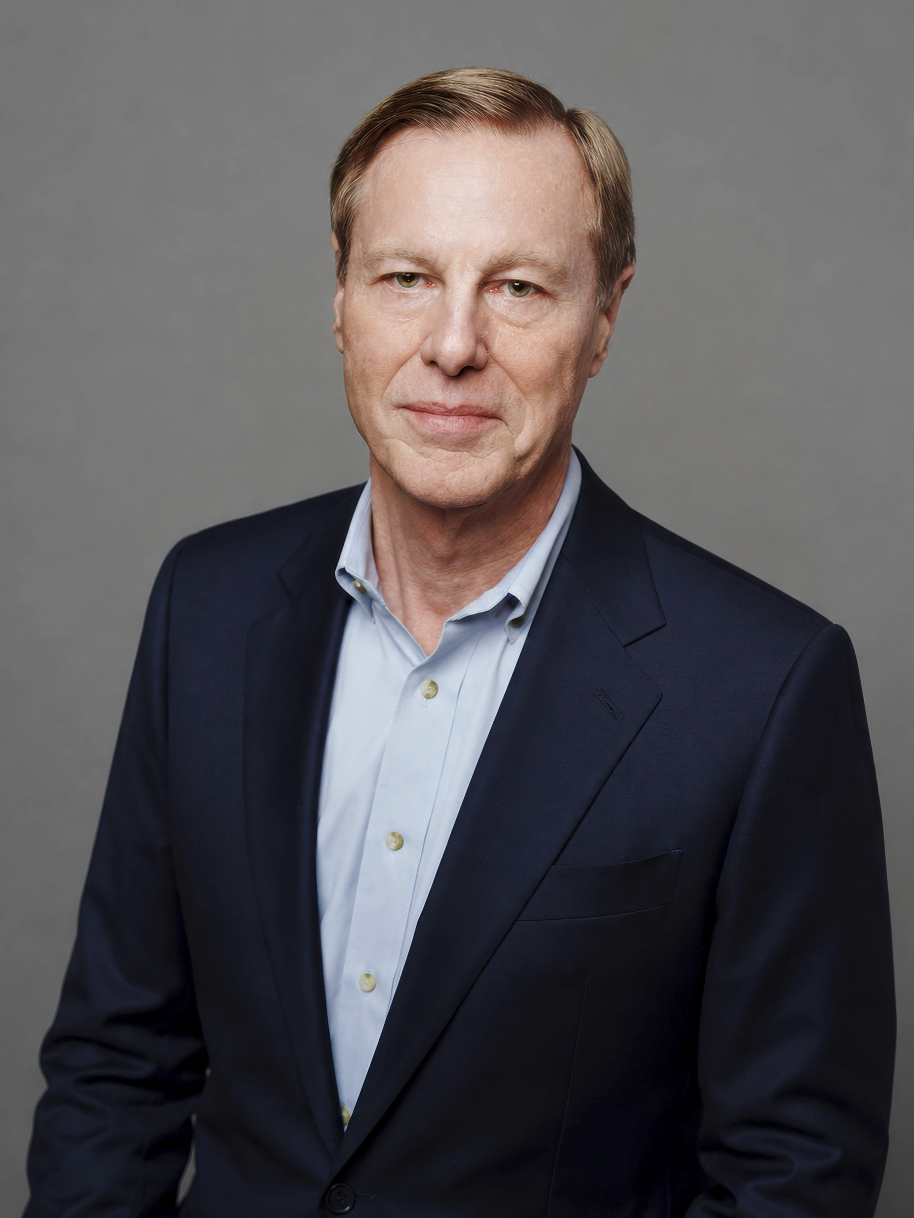 A middle-aged man with short light brown hair, dressed in a navy blue suit and light blue shirt, stands against a plain gray background for professional corporate headshots, looking at the camera with a neutral expression.