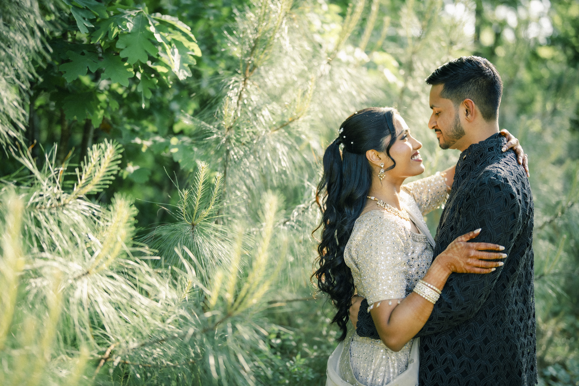 A couple dressed in elegant traditional attire, smiling and embracing each other in a lush green outdoor setting with sunlight filtering through the trees.
