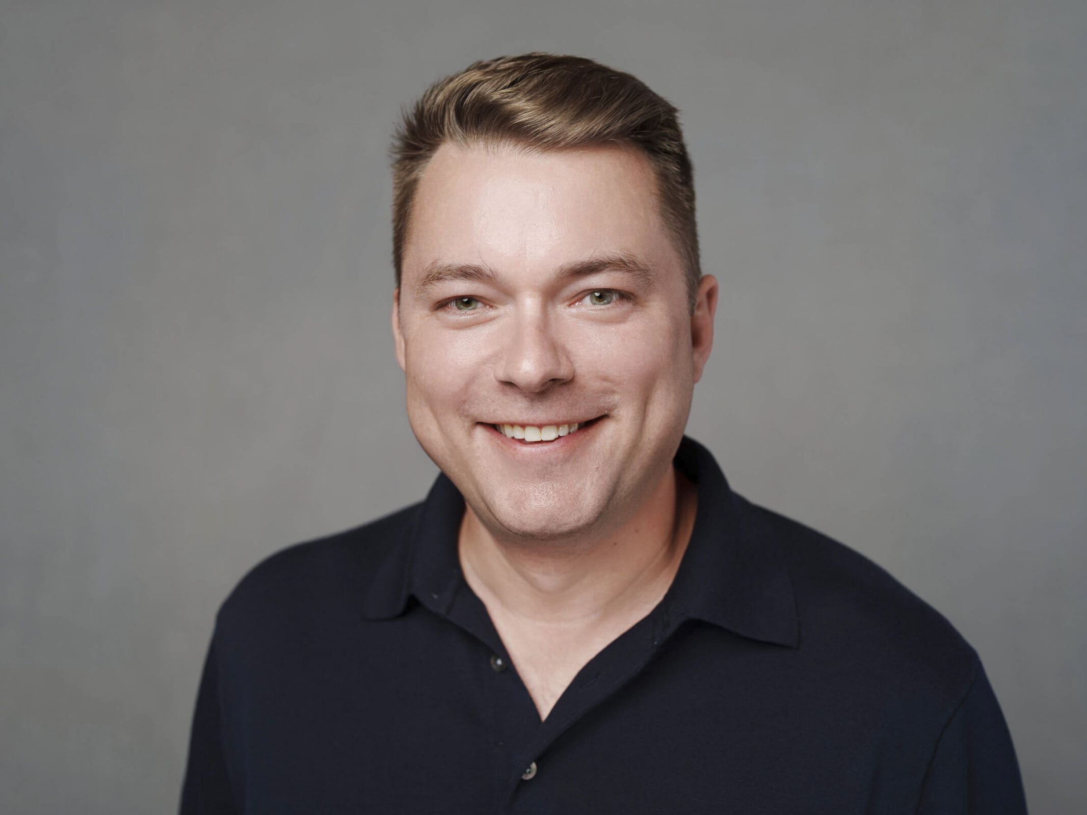A man with short light brown hair, wearing a dark navy collared shirt, smiles at the camera against a plain gray background, perfect for corporate headshots at The Umstead Hotel.