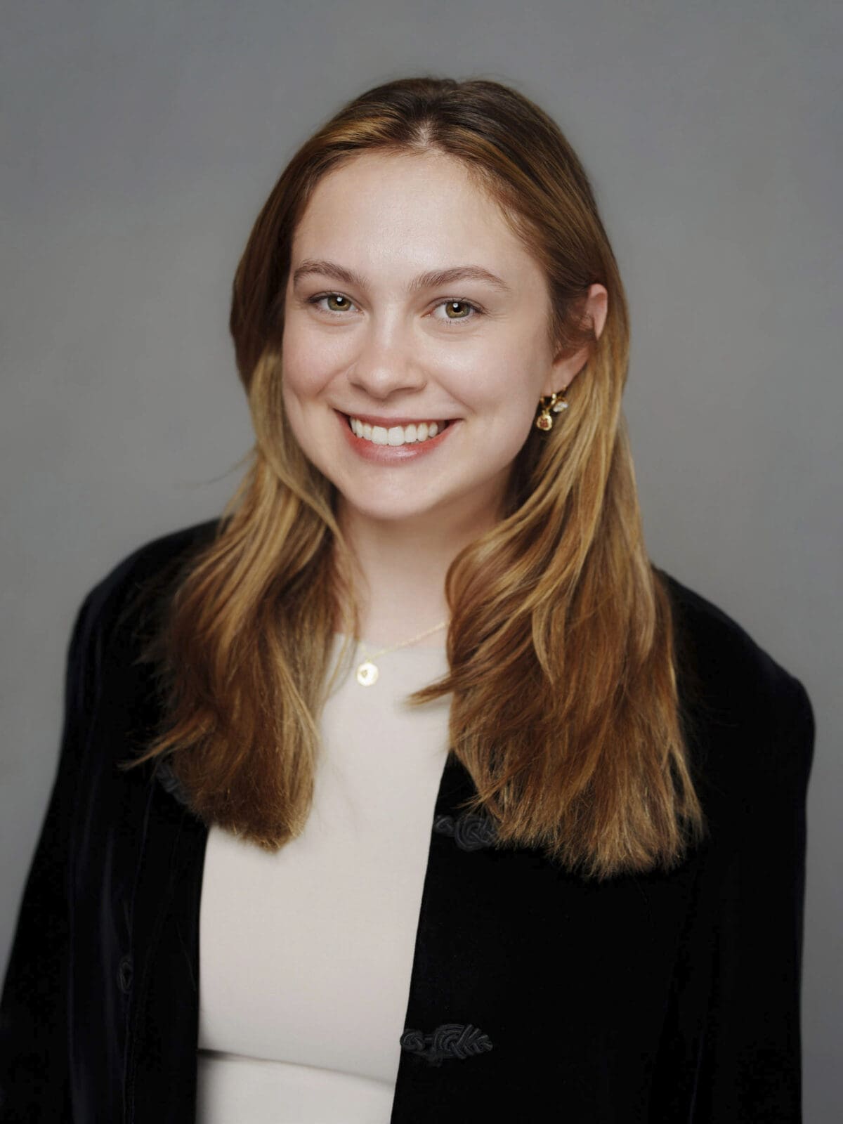 A young woman with long blonde hair is smiling at the camera in a classic corporate headshot. She is wearing gold earrings, a necklace, a cream-colored top, and a black jacket. The background is plain and gray.