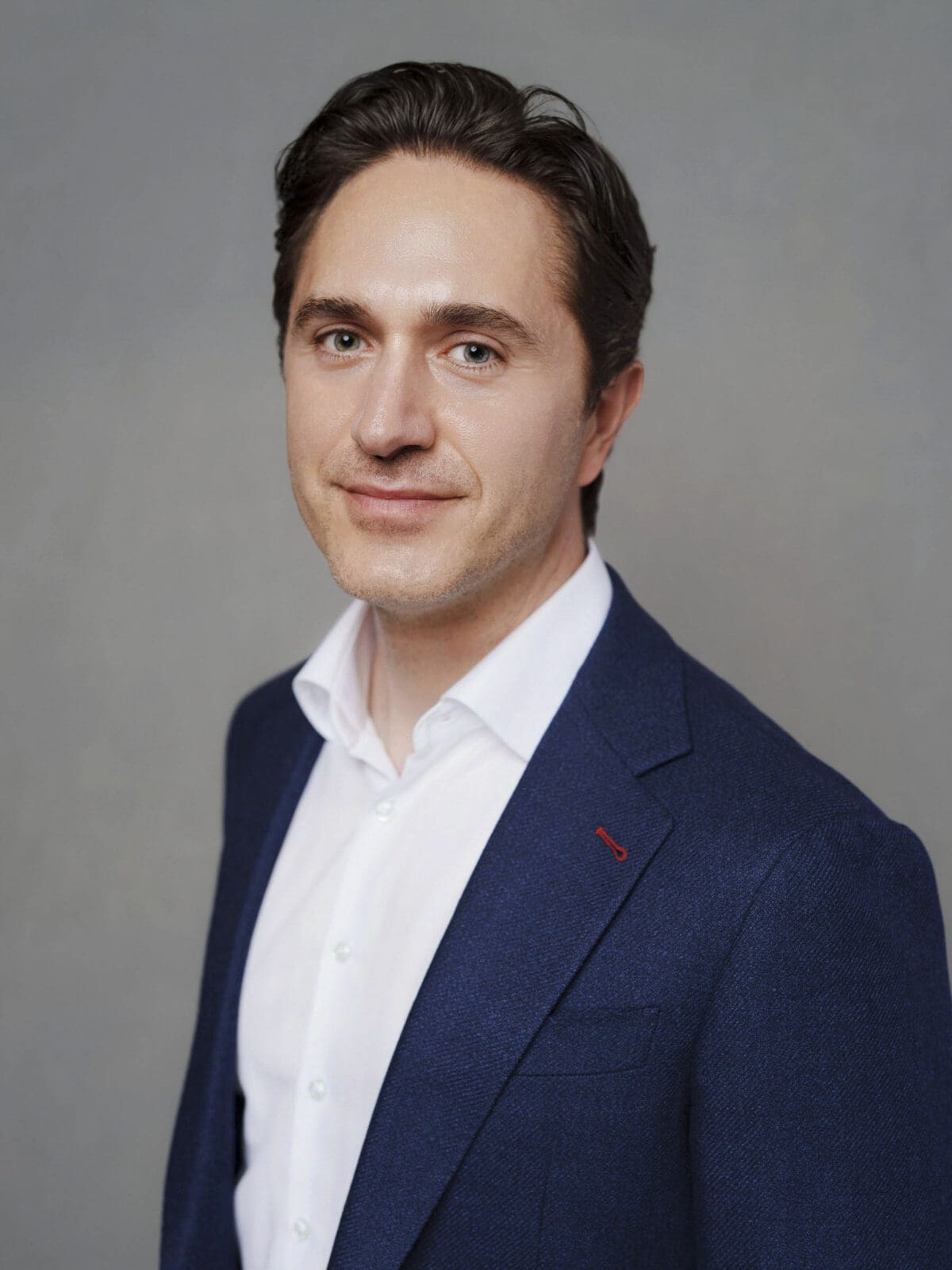 A man with short dark hair, wearing a white dress shirt and a blue blazer, smiles slightly while standing against a plain gray background—an ideal look for corporate headshots at The Umstead Hotel.
