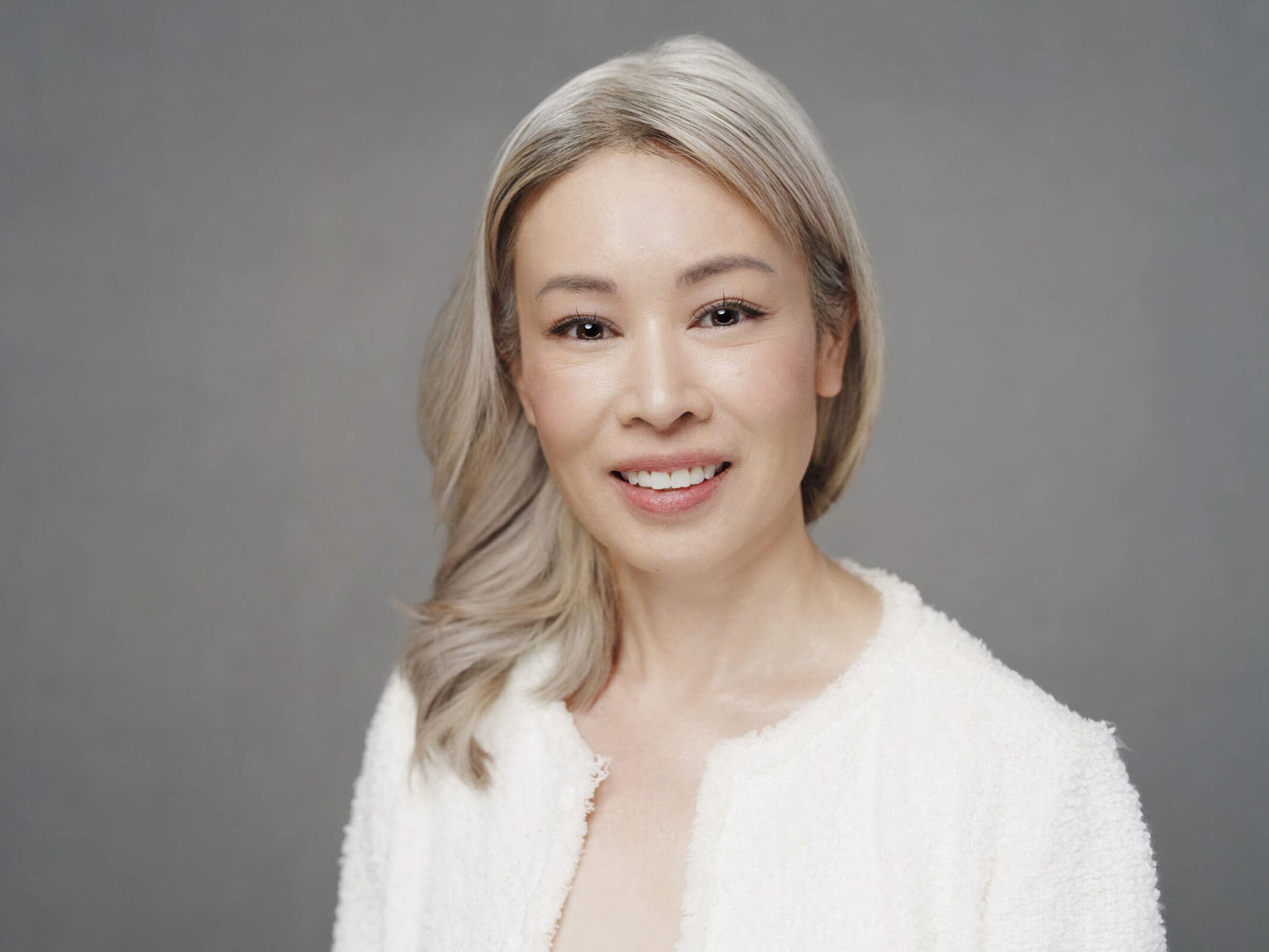 A woman with long, light gray hair smiles at the camera in a classic corporate headshot. She wears a white textured jacket and is posed against a plain gray background.