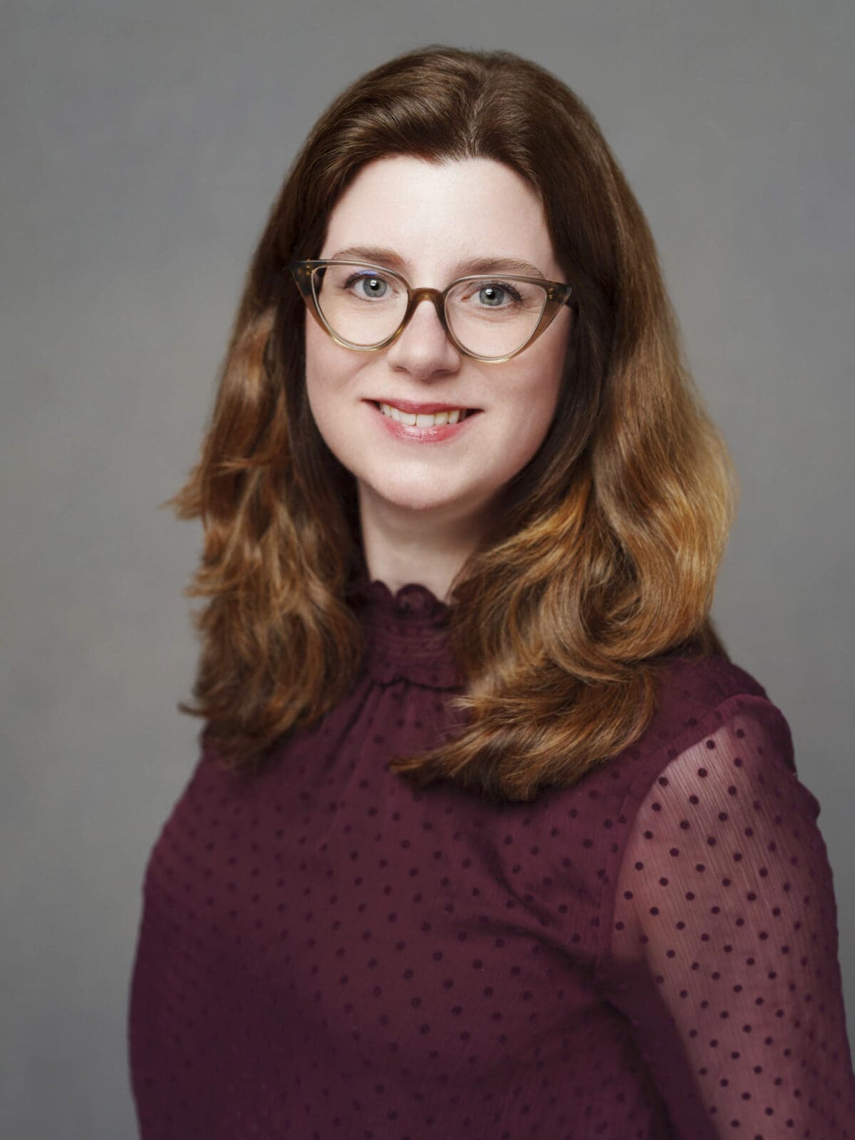 A woman with long, wavy brown hair and glasses smiles at the camera in a professional corporate headshot. She wears a burgundy, sheer polka-dot blouse against a plain gray background, perfect for The Umstead Hotel or Macrae’s executive profiles.