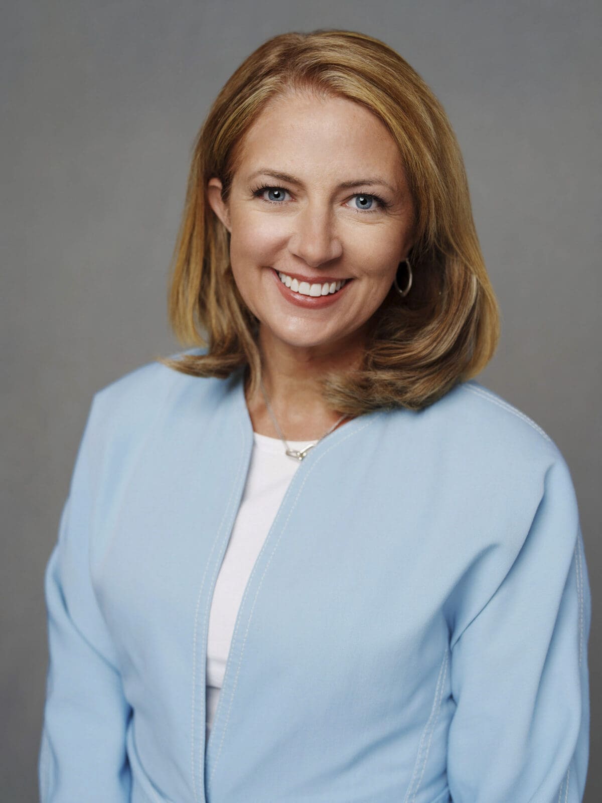 A professional corporate headshot of a woman with shoulder-length light brown hair wearing a light blue blazer over a white top, smiling warmly against a plain gray background at The Umstead Hotel & Spa.