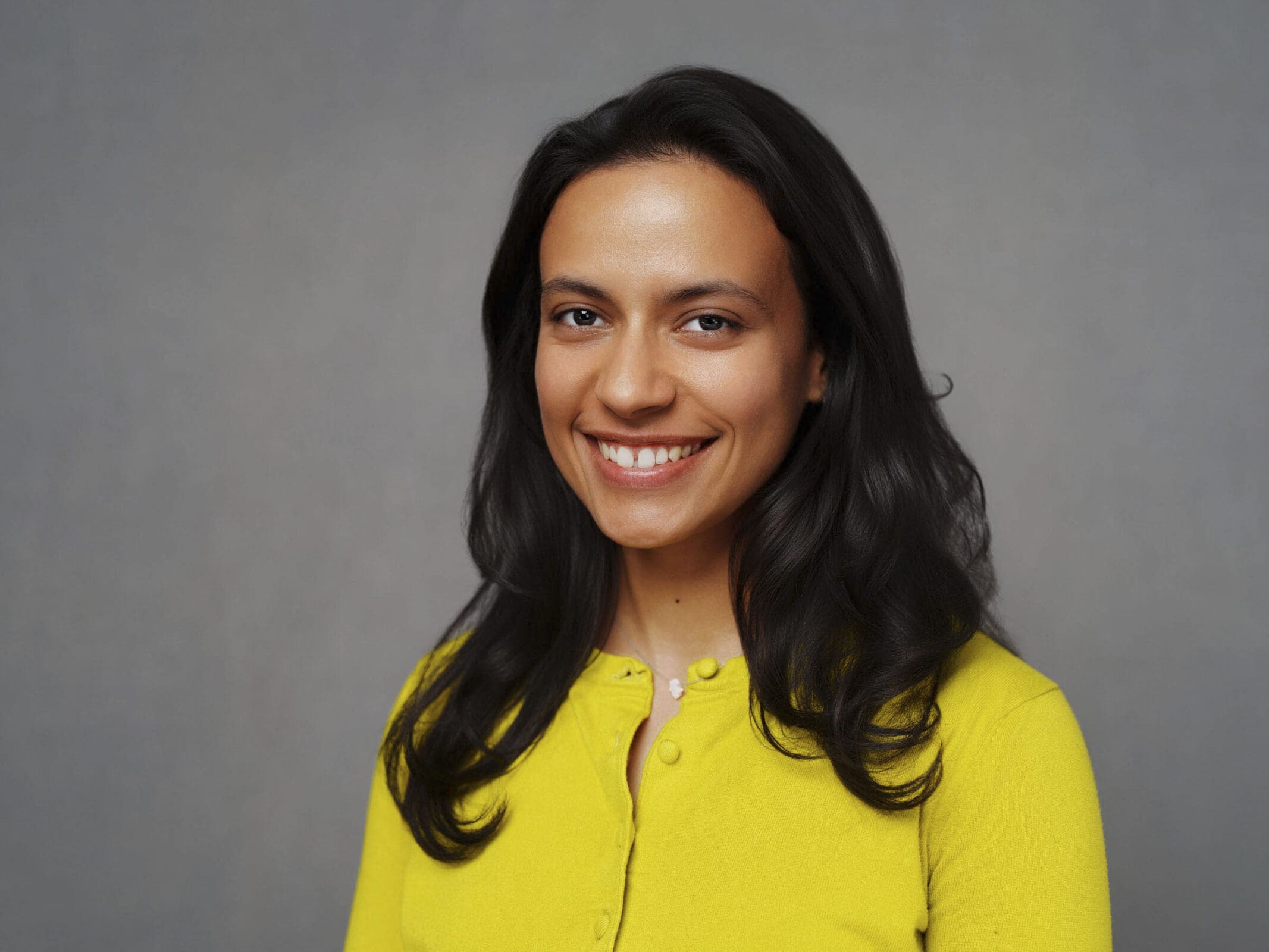 A woman with long dark hair smiles at the camera, wearing a bright yellow top against a plain gray background—an elegant example of corporate headshots inspired by Macrae’s style.