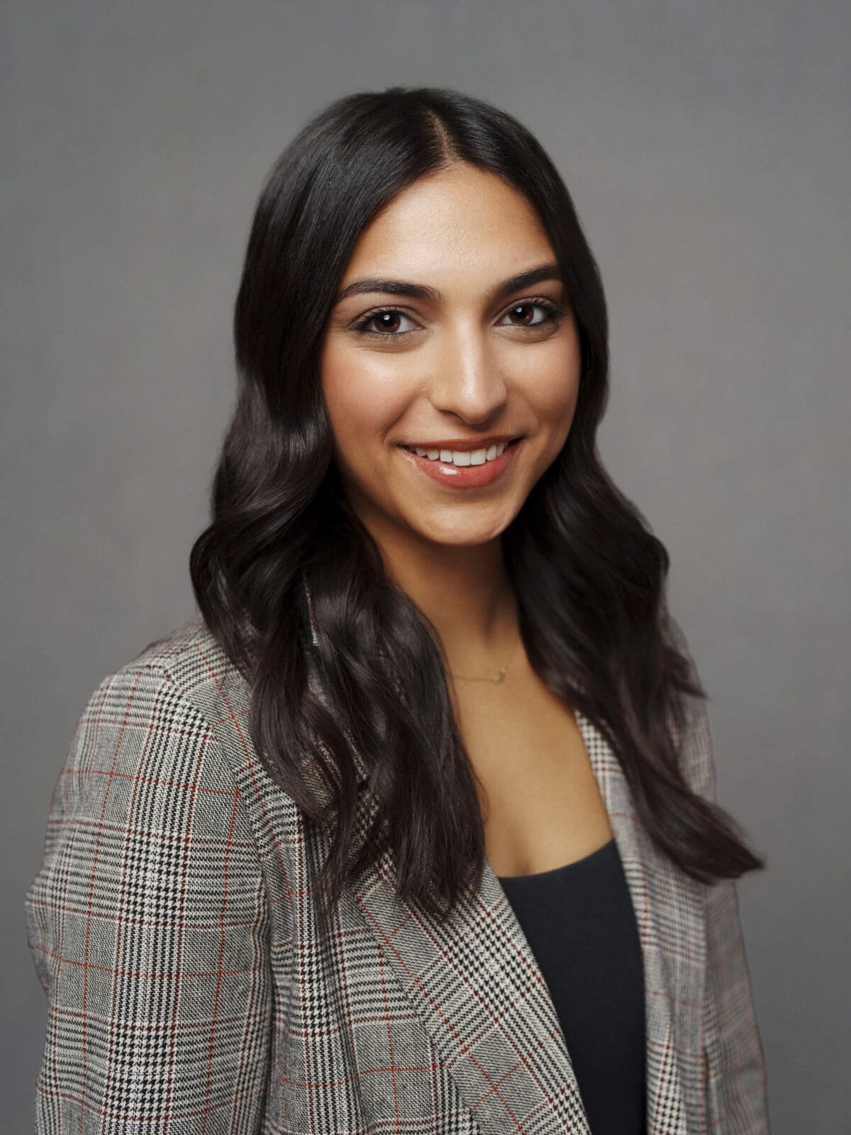 A woman with long, wavy dark hair smiles at the camera in a corporate headshot. She wears a plaid blazer over a black top, standing in front of a plain gray background. Captured by Macrae for The Umstead Hotel & Spa.