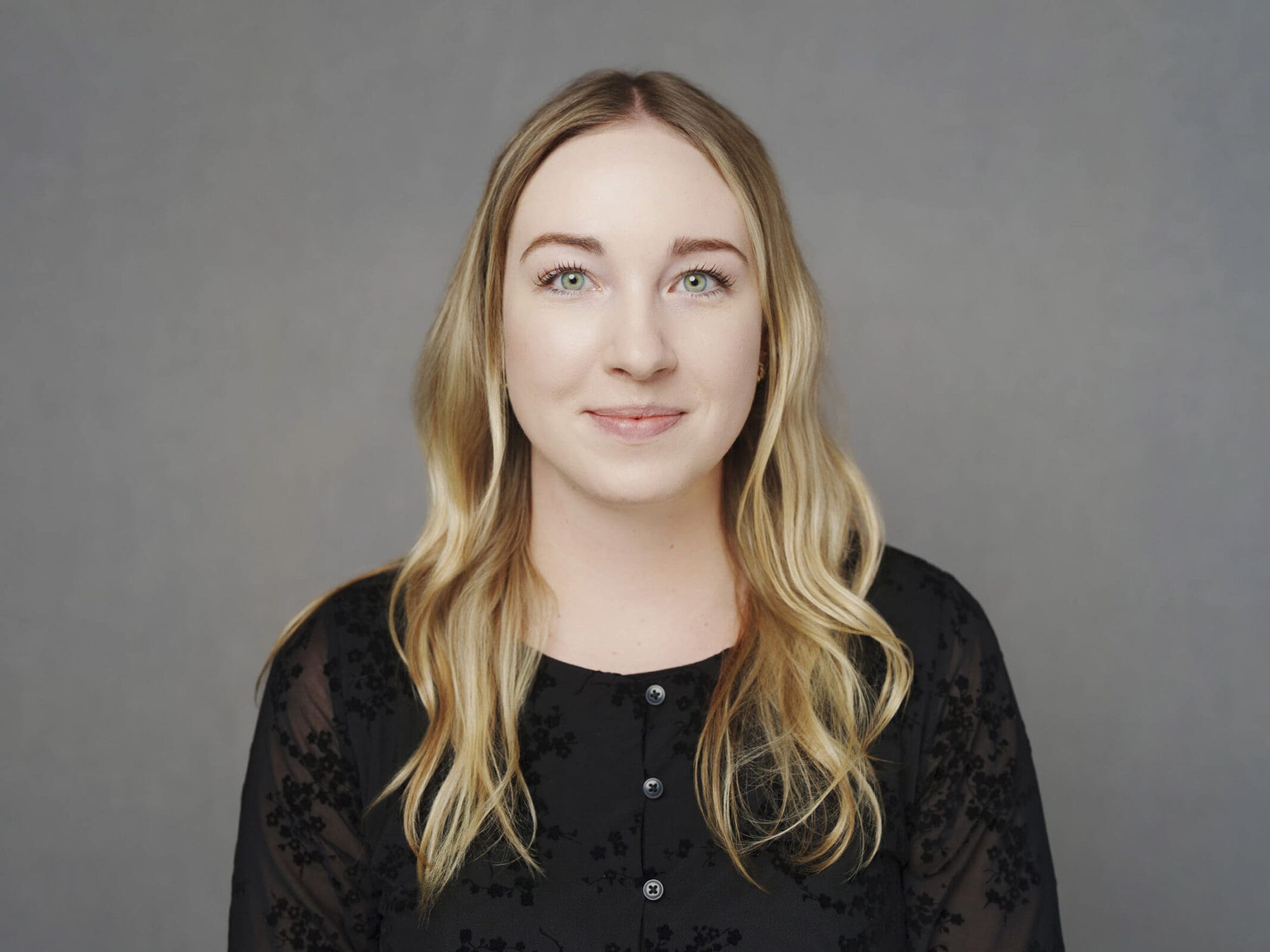 A woman with long blonde hair and green eyes, wearing a black top with buttons and sheer sleeves, poses for corporate headshots against a plain gray background, looking directly at the camera with a gentle expression.