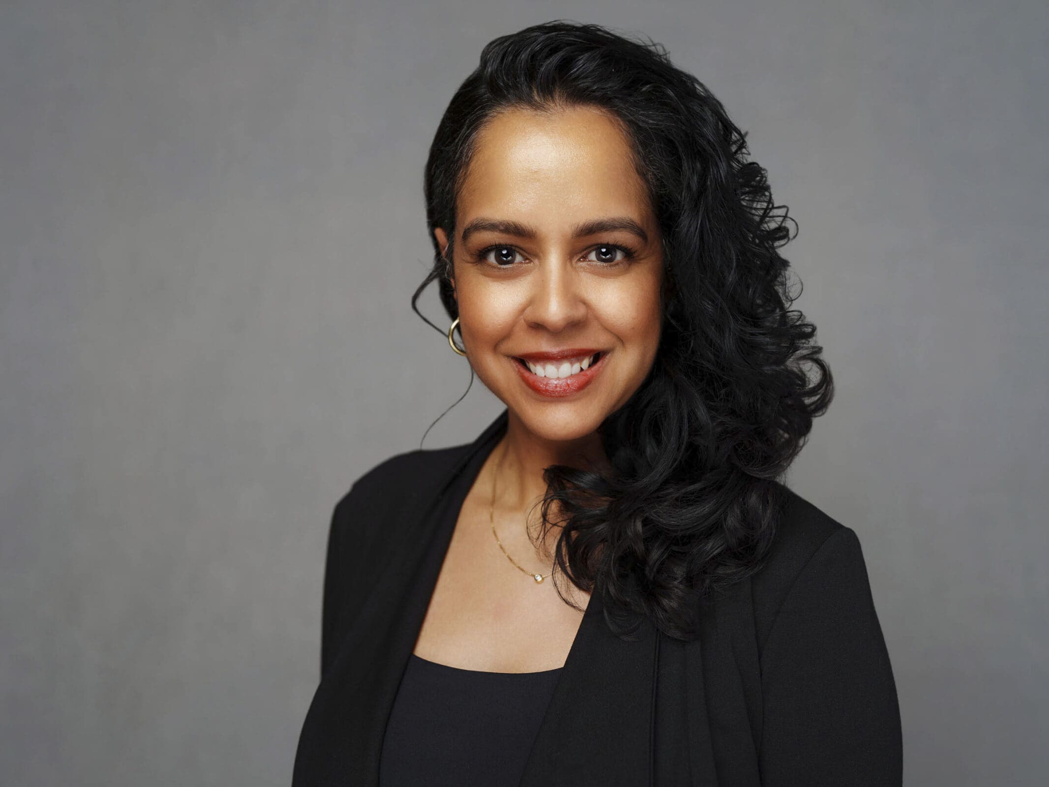 A woman with long, curly dark hair wearing a sleek black blazer smiles directly at the camera, an ideal example of confident corporate headshots against a classic gray background.