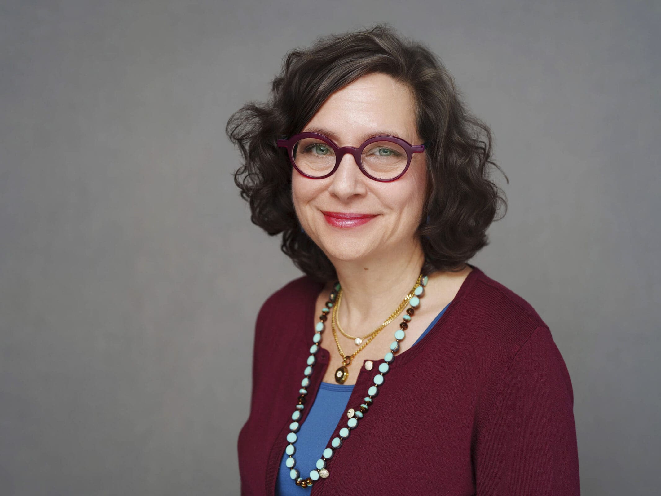 A woman with wavy brown hair wearing glasses with red frames, a burgundy top, a blue shirt underneath, and layered necklaces stands smiling in front of a plain gray background, perfect for corporate headshots at The Umstead Hotel.
