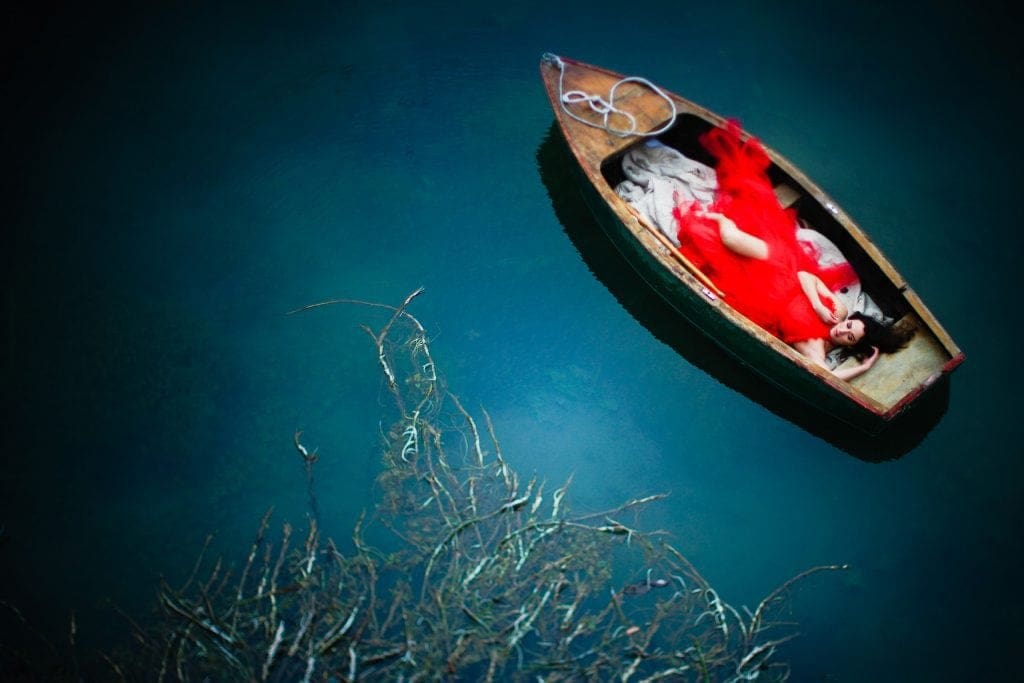 A woman in a flowing red dress lies in a wooden rowboat floating on calm blue-green water near Raleigh, with branches visible beneath the surface in the lower part of the image.