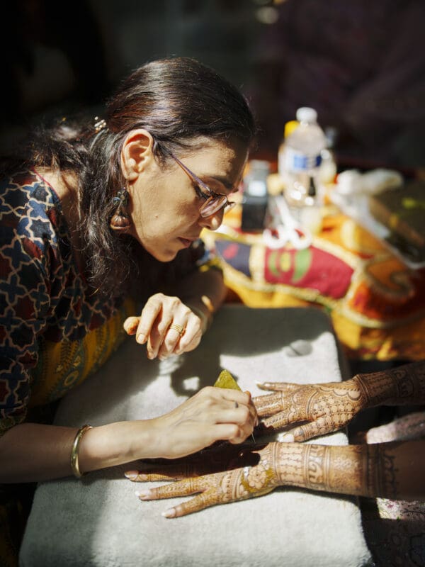 A woman with glasses carefully applies intricate henna designs to another person's hands, which rest on a towel. The brightly lit scene, filled with colorful fabrics and a water bottle, captures the artistry of a vibrant Raleigh Indian wedding.